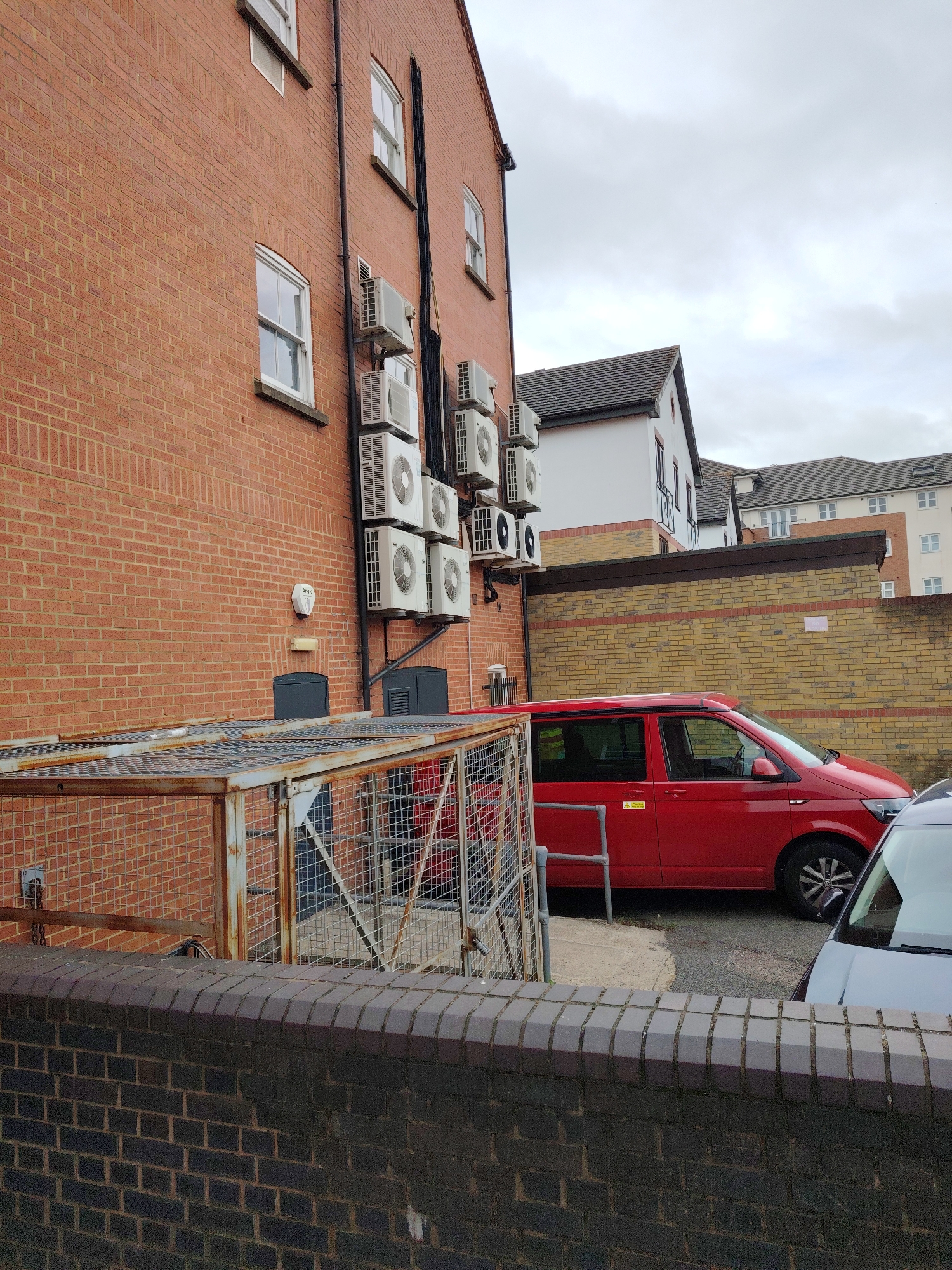 This image depicts a residential or commercial area in Peterborough, United Kingdom, under an overcast sky. The foreground features a low wall constructed from dark grey-brown bricks with a rounded coping. Behind this wall, a large, rusted metal cage structure with a mesh top and sides is visible, its frame appearing galvanized but aged.
To the left, a multi-story building constructed from reddish-brown brick dominates, featuring several white-framed sash windows with dark sills. Numerous white air conditioning condenser units are affixed to the brickwork, stacked vertically and horizontally, with black insulated piping running alongside them. A white security alarm box labeled "Angle" is mounted on the wall to the left of the AC units, above a dark recessed access panel.
In the midground, a red Volkswagen Transporter van with a pop-top roof is parked, oriented towards the right. A yellow warning sticker is affixed to its lower driver's side door. The front section of a dark grey or silver vehicle is partially visible in the immediate right foreground, parked adjacent to the red van. Behind the red van, a low wall constructed from pale yellow bricks features a distinctive horizontal stripe of darker reddish-brown bricks.
Beyond this wall, additional residential buildings are visible, characterized by white facades and dark-tiled pitched roofs, set against a uniformly cloudy, grey sky. No individuals are present in the scene.
