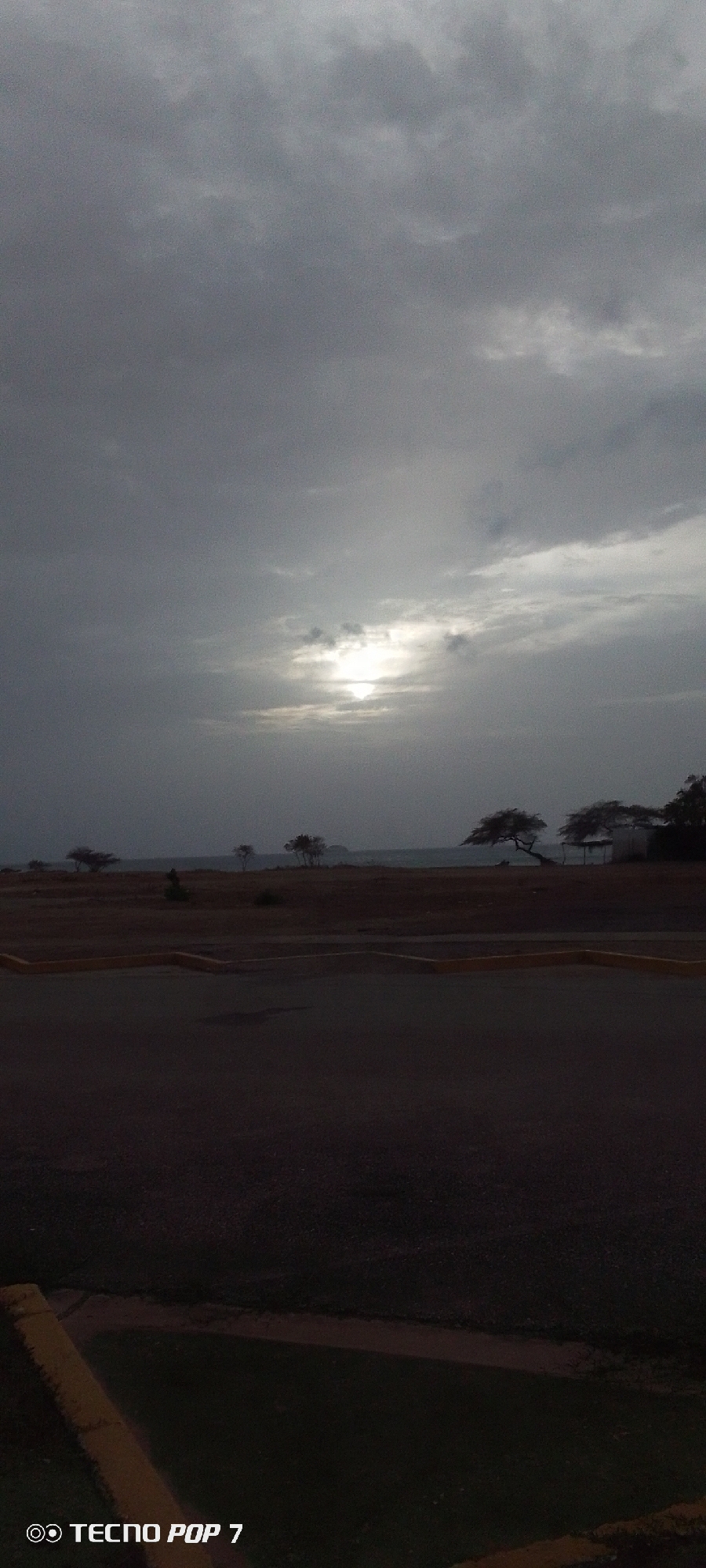 This photo, likely taken in Pedro González, Venezuela, shows a landscape under a heavy, overcast sky. The sun is faintly visible through the clouds, positioned near the center of the frame. The foreground features a dark, paved area, possibly a parking lot, with a section of grass and a yellow curb visible near the bottom. In the middle ground, there is a grassy, sandy area leading towards what appears to be a shoreline. Several trees dot the landscape along the shoreline, silhouetted against the sky. The "TECNO POP 7" watermark is visible in the bottom left corner.
