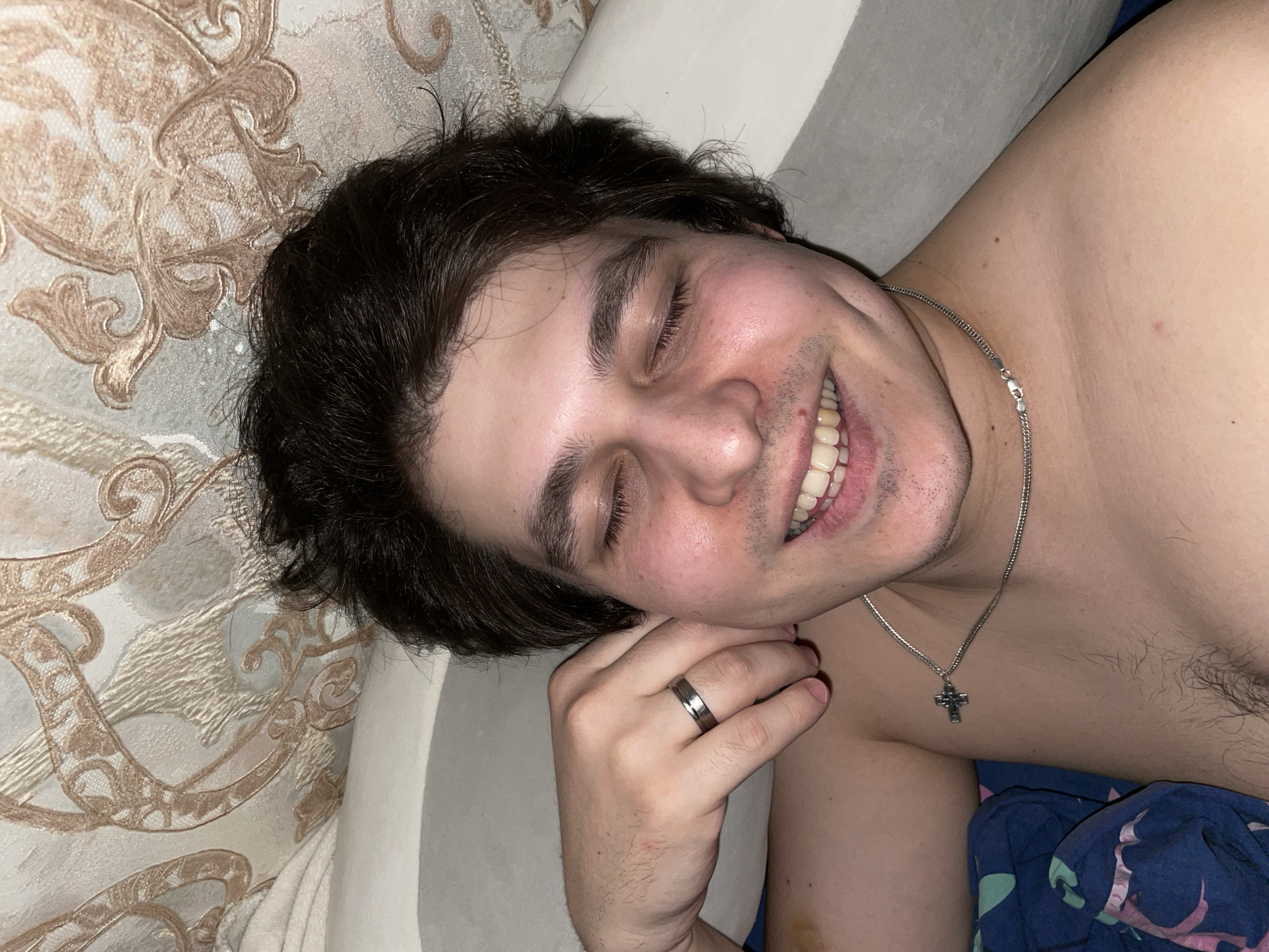 A man with dark hair is lying on a bed with his eyes closed and a smile on his face. He is wearing a silver ring on his left ring finger and a silver cross necklace. He is partially undressed, with a blue patterned sheet visible. The background includes a textured wall or headboard. The location is unknown and cannot be determined from the image.