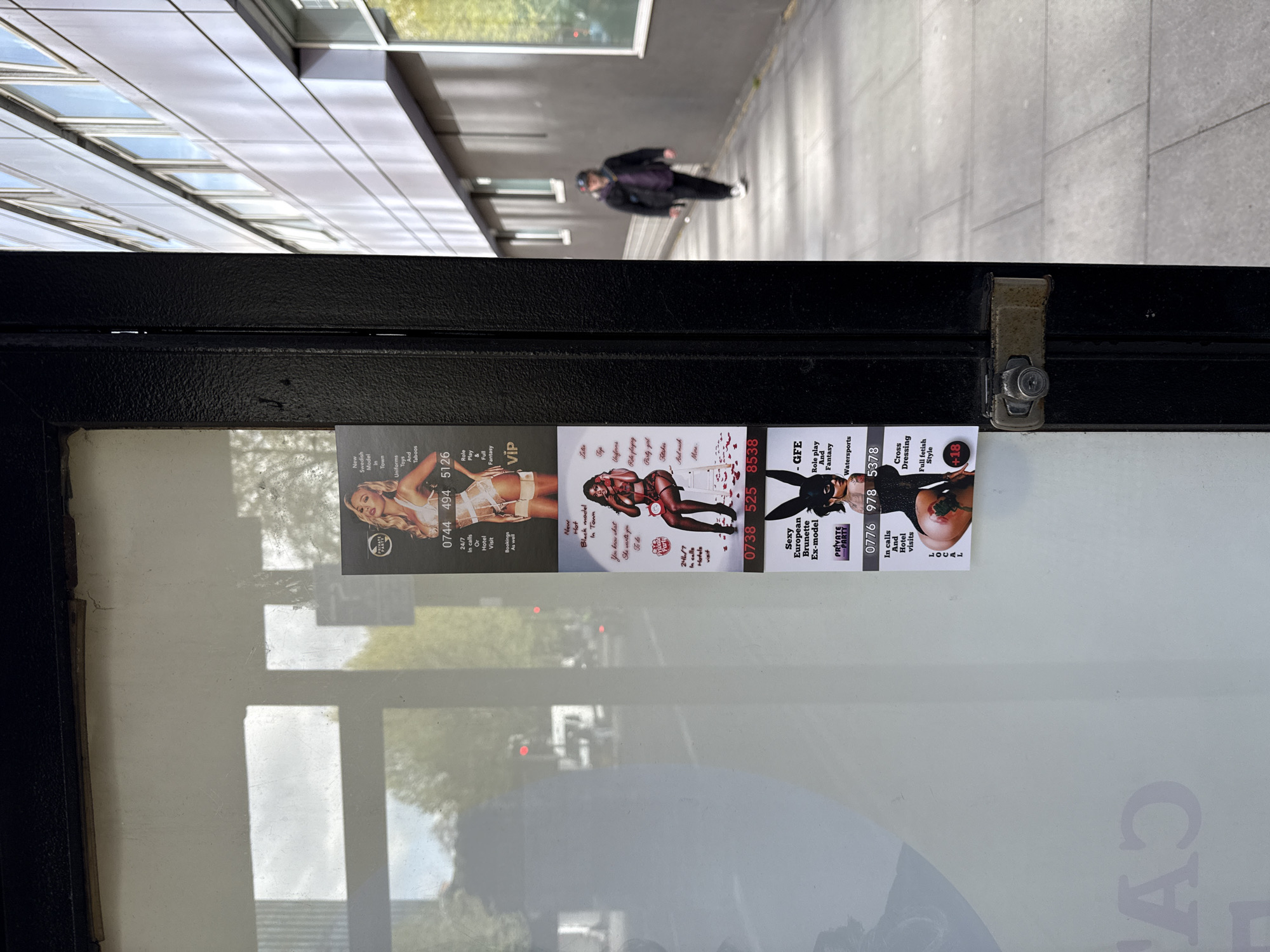 The image shows three advertisements adhered to the inside of a bus stop in a city that appears to be London, United Kingdom. These advertisements feature photos of women in lingerie and suggestive outfits. The ads include contact information. A person is walking in the background on a paved sidewalk. The bus stop is made of black metal and glass.