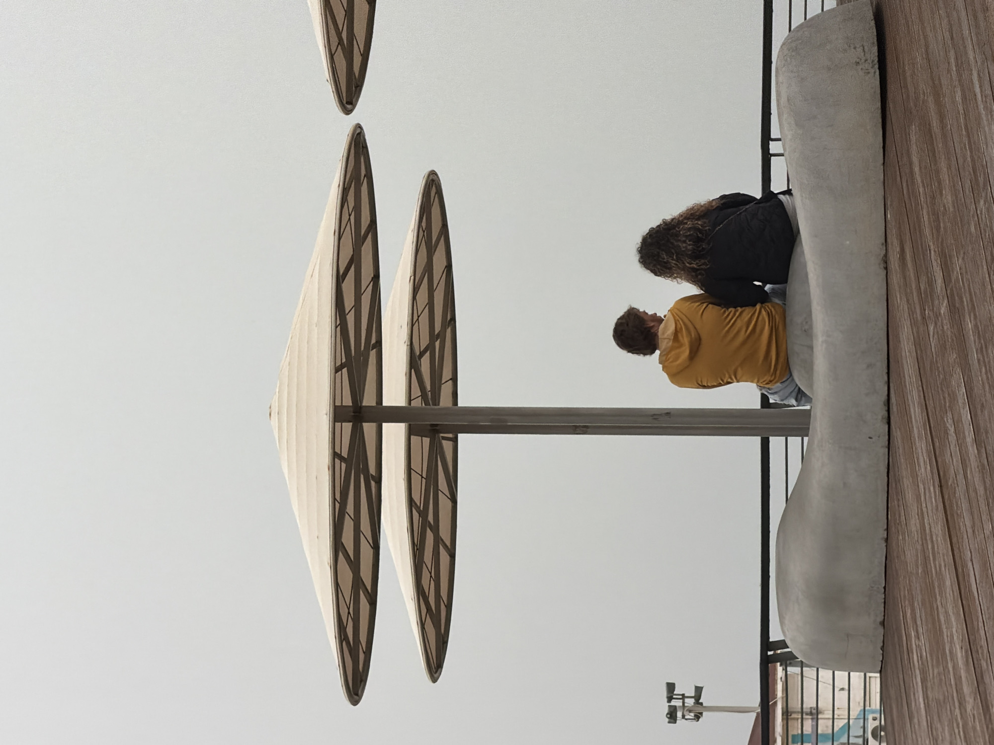 A low-angle photograph shows a couple sitting on a concrete bench in Tel-Aviv, Israel. The view is from below, looking up towards the sky, and the image appears to be rotated 90 degrees clockwise. The couple, one wearing a yellow shirt and the other a black jacket, is sitting close together. Above them are architectural sunshades that look like inverted cones with a grid pattern. A tall, grey pole supports the sunshades. To the right of the couple is a wooden deck surface that transitions into a concrete structure, acting as the bench seating. In the lower right corner, a security camera and a portion of a building are visible beyond the bench, suggesting a commercial area. The sky is an overcast grey, filling most of the background.