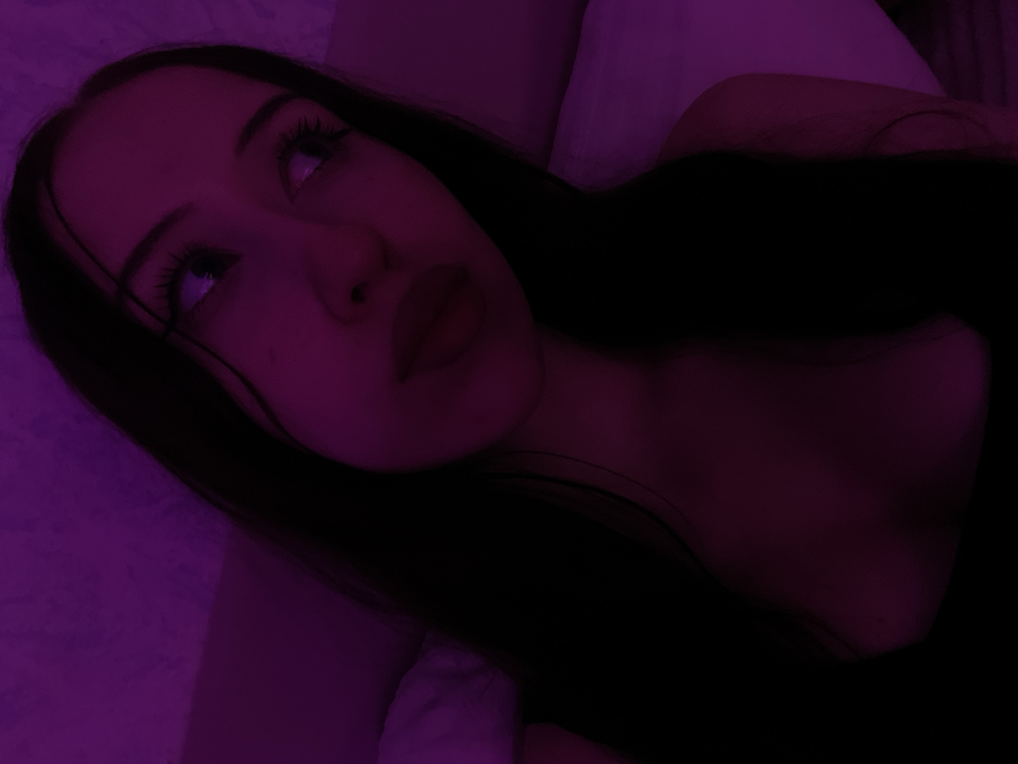 The image shows a close-up of a young woman with long, dark hair, illuminated by purple light. She is looking off to her right. Her eyes are prominent, with visible eyelashes, and her lips are slightly parted. She is wearing a dark-colored top, the neckline of which is visible. The background appears to be a textured surface, possibly a wall or bedding, also cast in the same purple hue. The scene suggests an indoor setting, likely a bedroom, given the soft lighting and pillows visible on the right side of the frame. The image does not contain any elements that would allow for the identification of a specific location within Votkinsk, Russia.