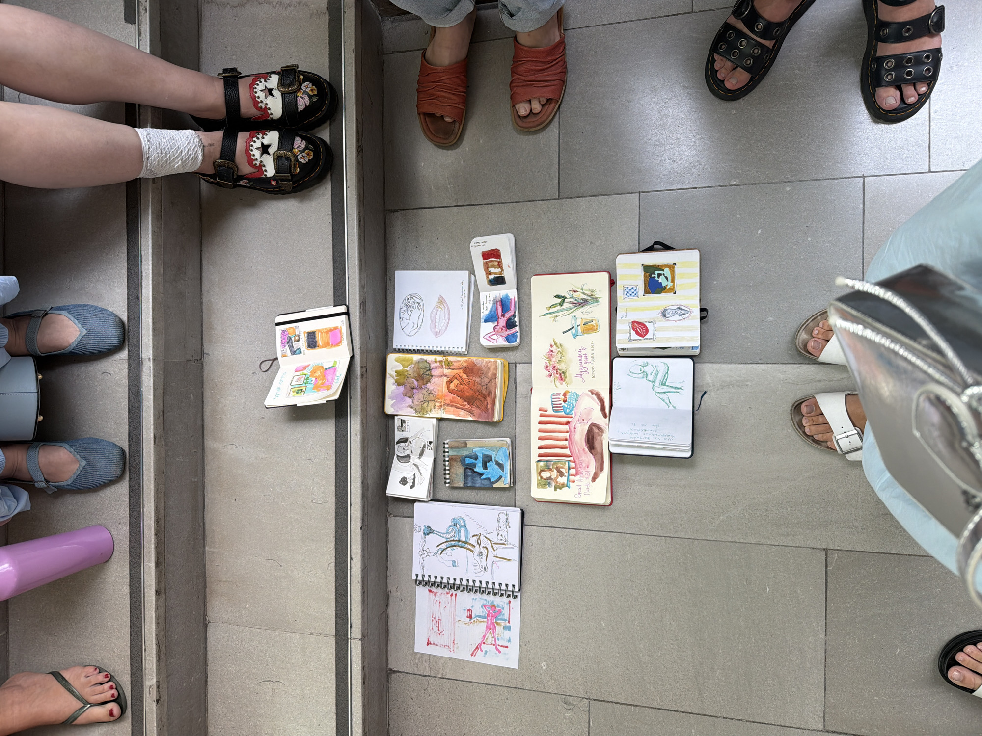 The image shows a top-down view of several people's feet and legs, along with a collection of sketchbooks laid out on a tiled floor. The setting appears to be an outdoor, public space, possibly a staircase or pathway, given the concrete steps and the presence of a railing. The lighting suggests it is daytime, with no strong shadows indicating a particular time of day.
The subjects are diverse in their footwear. One person is wearing black Mary Jane style shoes with floral embroidery and buckles. Another is wearing red, crinkled leather slide sandals. A third person has on black sandals with multiple buckles and eyelets, and a fourth is wearing white Birkenstock-style sandals. Two individuals are wearing flat, light blue slip-on shoes, and another is wearing dark grey flip-flops.
The sketchbooks are open and display various drawings and paintings. Some of the sketches are of human figures, while others appear to be still lifes or abstract compositions. The artwork ranges in style from simple line drawings to more detailed watercolor paintings. One sketchbook has text in what appears to be Cyrillic script.
The overall impression is one of a casual gathering, possibly an art class or a group of friends who are sketching together outdoors. There are no overt emotional cues, but the relaxed postures and the shared activity suggest a friendly and creative atmosphere. The location context provided is Tel-Aviv, Israel.