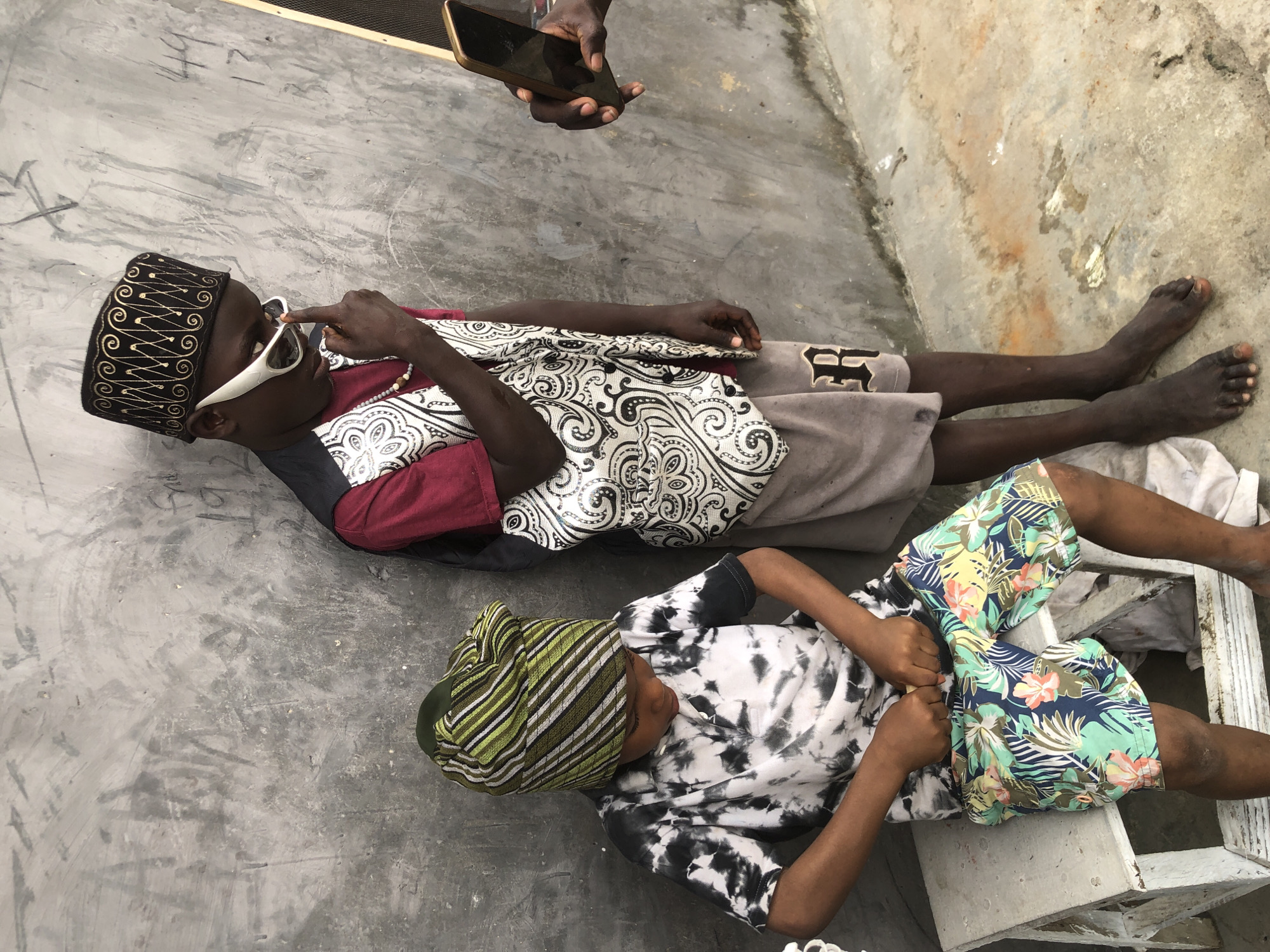 Two young Black males are depicted in an outdoor setting against a textured gray concrete wall. The male on the left, seated on a white wooden bench, wears a tie-dye t-shirt with black and white splotches and floral-patterned shorts. He is looking down with his hands near his waist. The male on the right stands and wears a maroon shirt, a patterned vest, and gray shorts. He has a black kufi cap on his head and white-rimmed sunglasses. His right hand is raised to his chin, touching his lips with his index finger. To the right of the standing male, a hand holds a smartphone, suggesting a photo is being taken. The ground is concrete, with some debris and discoloration. The overall scene appears to be in Ado, Nigeria.
