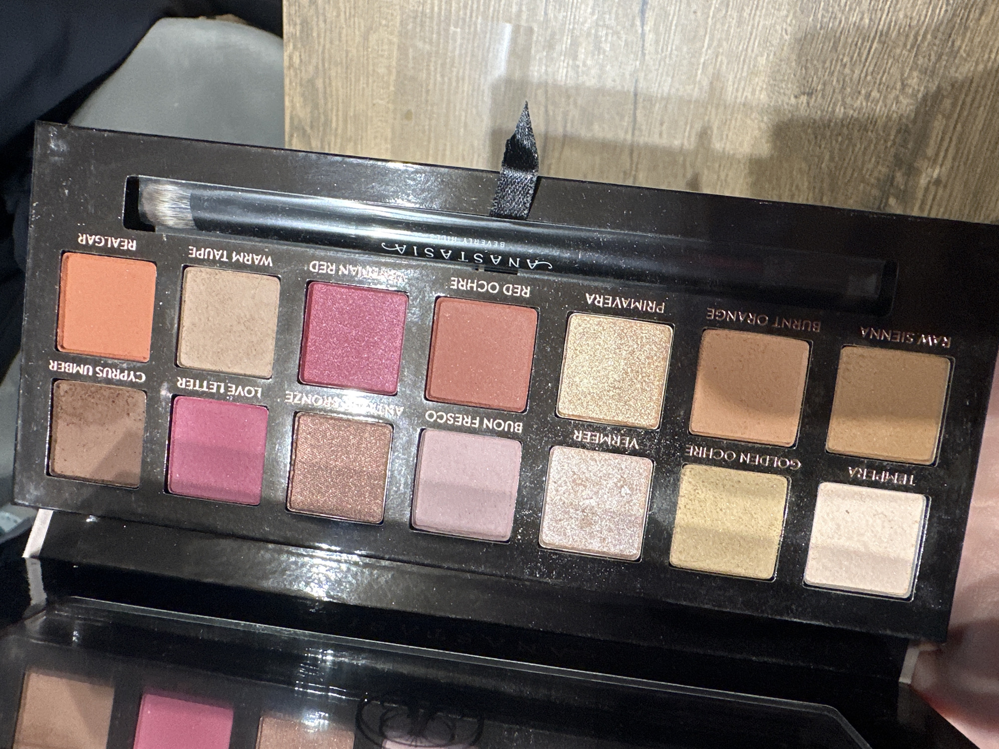The image shows an Anastasia Beverly Hills eyeshadow palette, specifically the Modern Renaissance palette, open and displaying its various colors. The palette includes shades such as "Realgar," "Warm Taupe," "Red Ochre," "Primavera," "Burnt Orange," "Raw Sienna," "Cyprus Umber," "Love Letter," "Bronze," "Buon Fresco," "Vermeer," "Golden Ochre," and "Tempera." A double-ended eyeshadow brush is present, sitting above the eyeshadows. The brand name "ANASTASIA BEVERLY HILLS" is printed in white on the plastic film over the brush. The palette has a black, matte finish. The environment appears to be indoors, near a wood surface. There is no information to determine that this photo was taken in Plovdiv, Bulgaria.