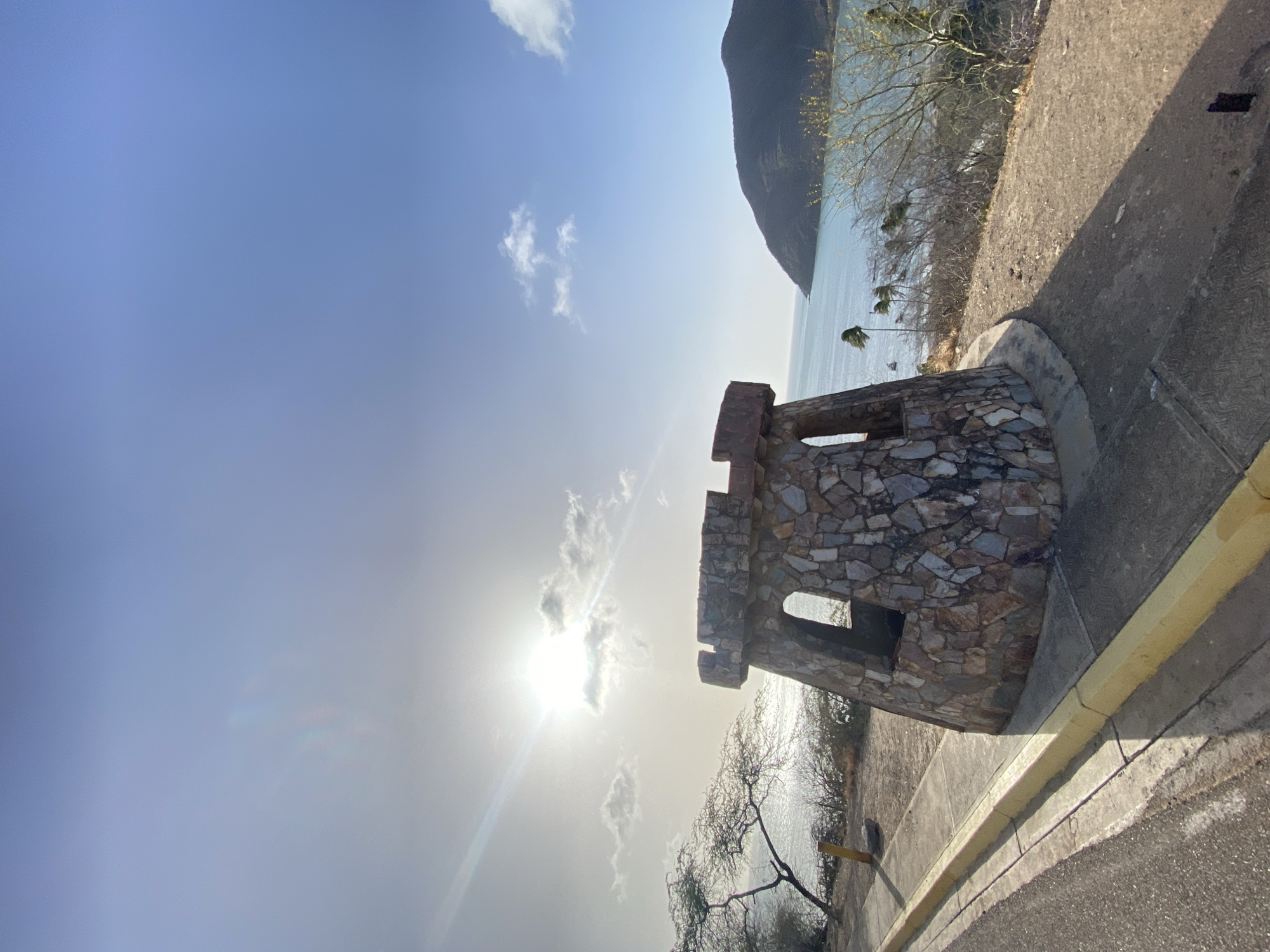 A stone tower-like structure stands next to a paved road with a yellow curb. Beyond the tower, the sea stretches towards a headland under a sky with scattered clouds. The sun shines brightly, positioned behind a cloud in the sky, creating a glare. Thin, bare trees line the edge of the cliff. The location is Juan Griego, Venezuela.