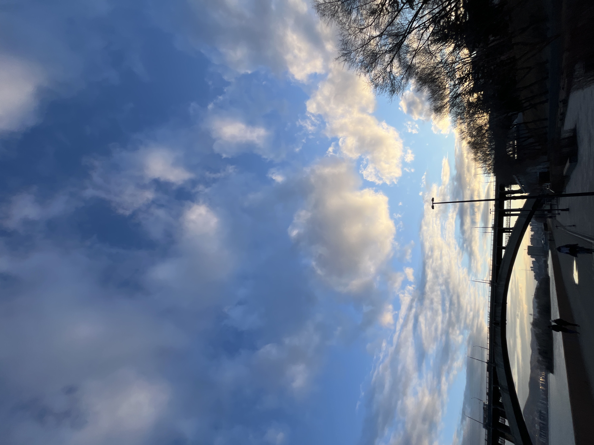 The image, oriented horizontally, captures an expansive outdoor scene featuring a prominent bridge spanning a wide body of water under a dynamic sky. The time of day appears to be late afternoon or early evening, evidenced by the low sun casting warm, golden light on the edges of scattered clouds against a deep blue sky.
A large, curved bridge, likely carrying a road or pedestrian path, dominates the lower part of the frame, equipped with several lampposts. Below it, a calm river or wide body of water reflects the sky's soft light. On the distant bank, a dense line of trees with bare branches forms a dark silhouette, suggesting late autumn or winter. Beyond these trees, a faint cityscape with indistinct buildings is visible on the horizon.
Several individuals are present on the bridge's walkway, appearing as dark, silhouetted figures walking or possibly running. The scene conveys a sense of peaceful serenity, enhanced by the interplay of light and shadow. The setting is Krasnoyarsk, Russia, likely along the Yenisei River. No visible text is present in the image.