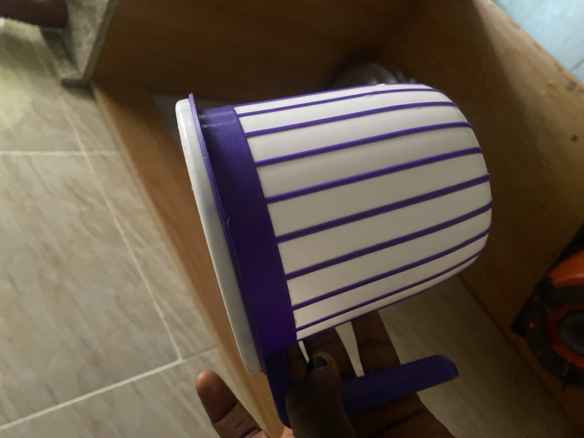 A hand holds a white and purple striped mug with a purple handle, positioned over a wooden countertop. The mug has a white lid. Behind the mug, a tiled wall is visible. The scene appears to be in a kitchen or bathroom area. The lighting suggests indoor illumination.