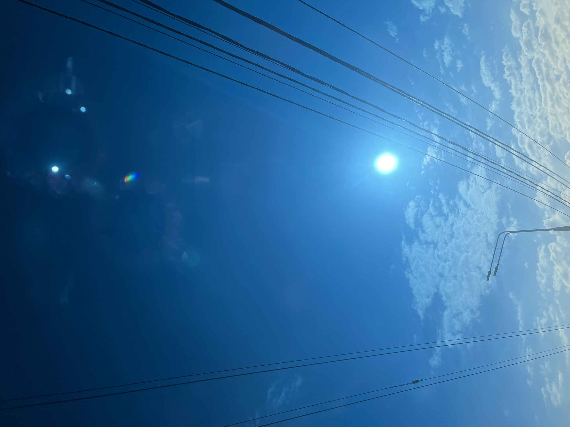 The image depicts a low-angle view of a sky with high-voltage power lines crisscrossing the frame. The sun is visible as a bright point of light, causing lens flare. Parts of a utility pole with streetlight fixtures are visible in the upper right corner. The sky is blue, with some streaky white clouds in the upper right. The location is likely Krasnodar, Russia, as that was specified in the instructions.
