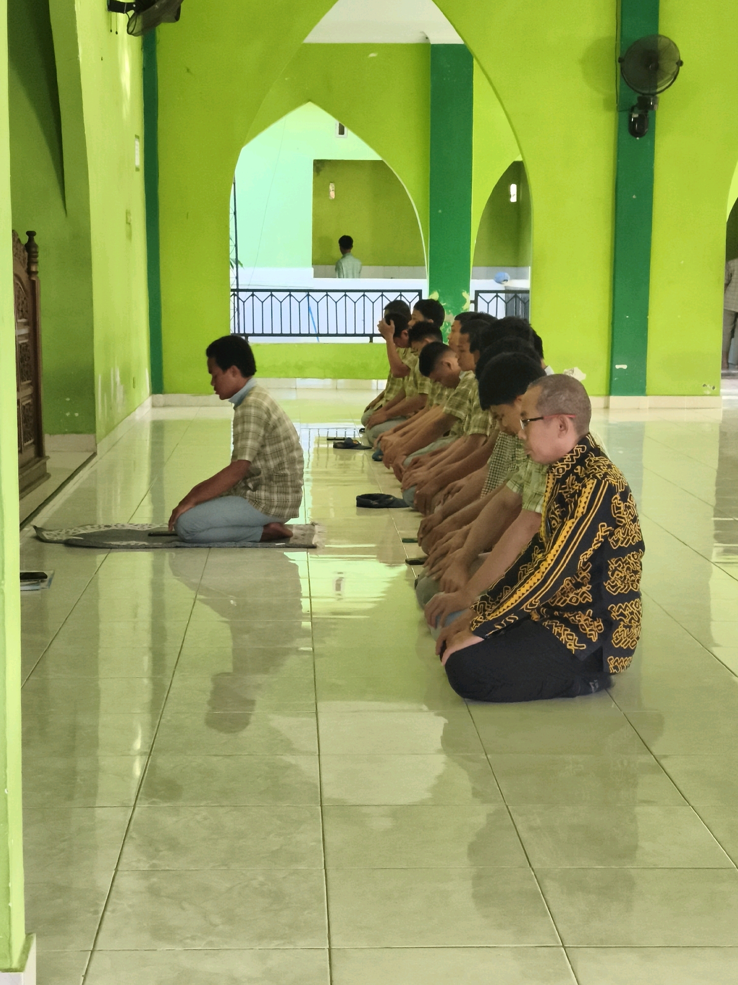 A group of young men and one older man are seated on the floor of a mosque in Makassar, Indonesia, in a prayer formation. The room is painted a vibrant lime green, with arched doorways leading to other sections of the building. The floor is tiled and highly reflective. The men are all facing the same direction, with their hands resting on their laps. The younger men are wearing similar light-colored shirts, while the older man is wearing a black shirt with a gold and yellow patterned design. A prayer mat is laid out in front of the first man on the left. In the background, through an archway, another individual can be seen standing. The scene appears to be one of quiet contemplation and religious observance.