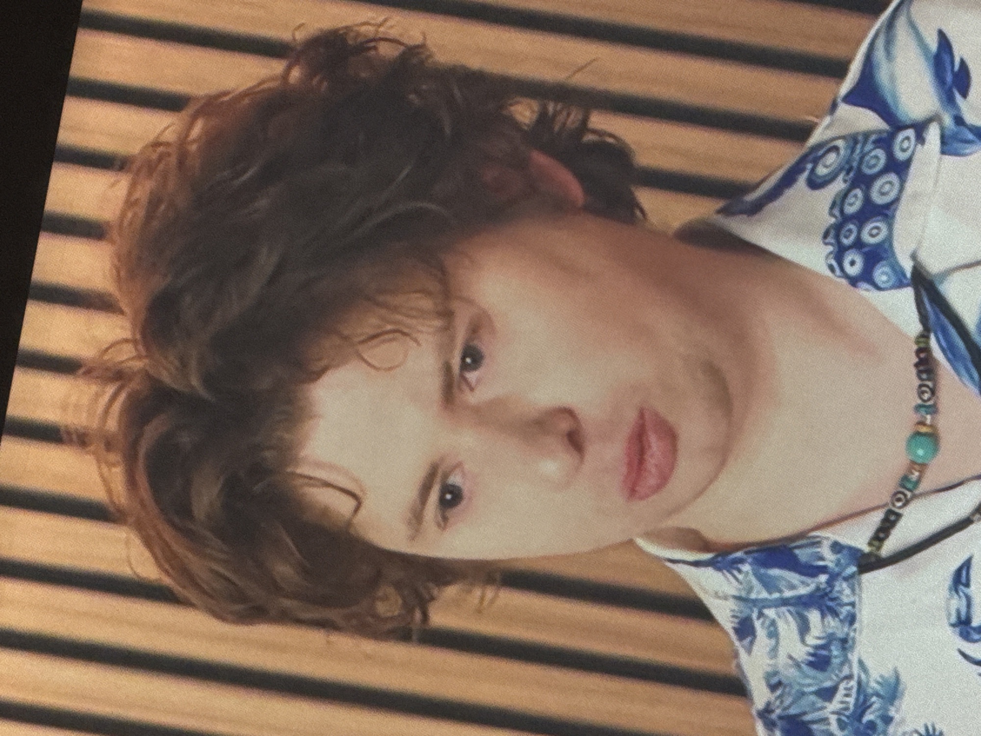 A medium shot shows a person with wavy, brown hair and fair skin. They are wearing a white shirt with a blue floral and tentacle pattern, and a beaded necklace with a turquoise pendant. The person's mouth is slightly open, and their expression is neutral. The background consists of vertical wooden slats in shades of brown and black. The image is a photograph, likely taken indoors. There is no information to suggest the location is Saratov, Russia.