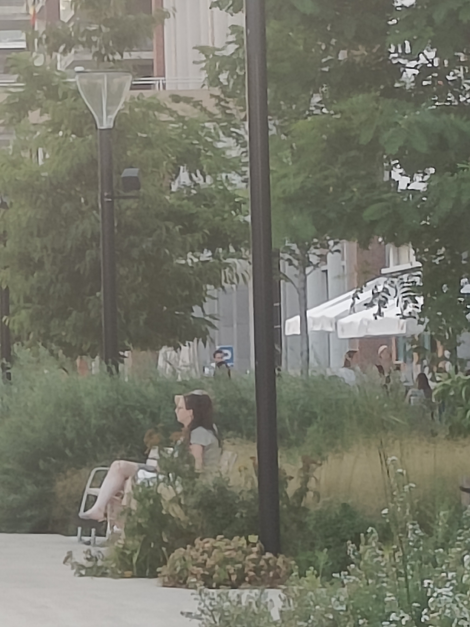 This is a medium-distance, eye-level shot of a park-like area with people seated on benches. The scene is characterized by lush greenery and trees in the background, with a building facade visible behind the foliage. A tall, dark pole stands prominently in the foreground, slightly to the right of the center. To the left of this pole, a street lamp with a translucent, bell-shaped top is visible, also on a dark pole. In the middle ground, a woman with dark hair is seated on a white chair, facing left, her legs crossed, and she appears to be looking to her left. She is wearing a light-colored shirt. Behind her, more people are visible, some sitting at tables under white awnings, suggesting an outdoor cafe or restaurant. The ground in the foreground is a light-colored, paved surface. The overall atmosphere suggests a pleasant, leisurely outdoor setting in an urban environment. The presence of the street lamp and the architectural style of the building suggest this scene is located in the city of Leuven, Belgium.