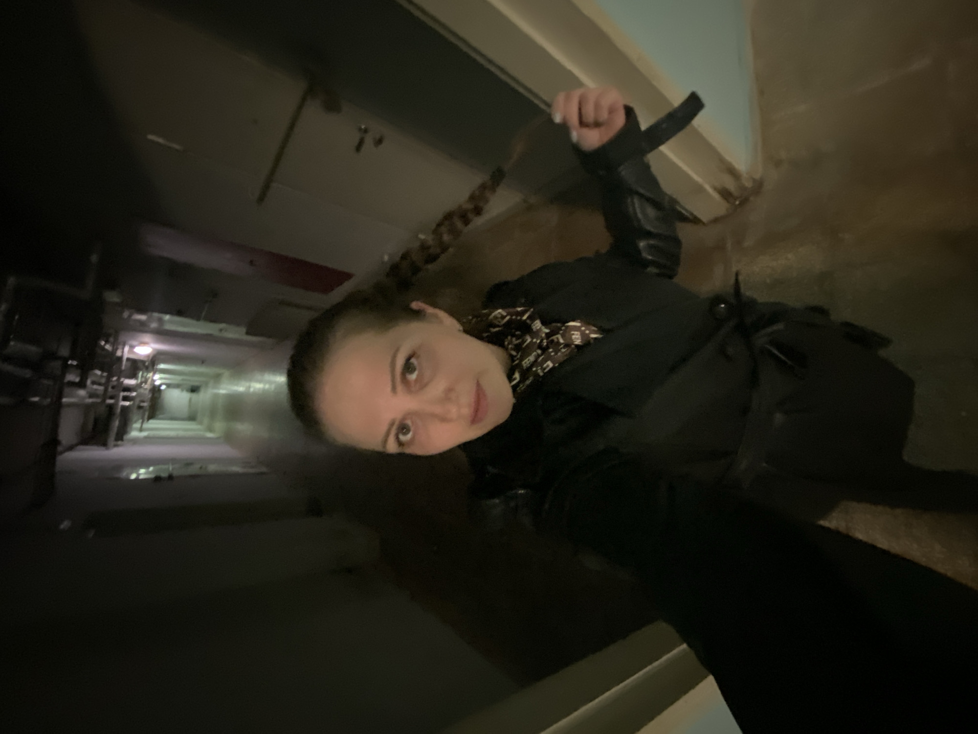 A person stands in a dim hallway, likely indoors, potentially in a building or facility in Марковский, Russia. The person has a braid and is wearing a dark-colored top or jacket. The hallway has a series of doors on the left, a wet floor with some puddles, and a light at the end of the hallway.