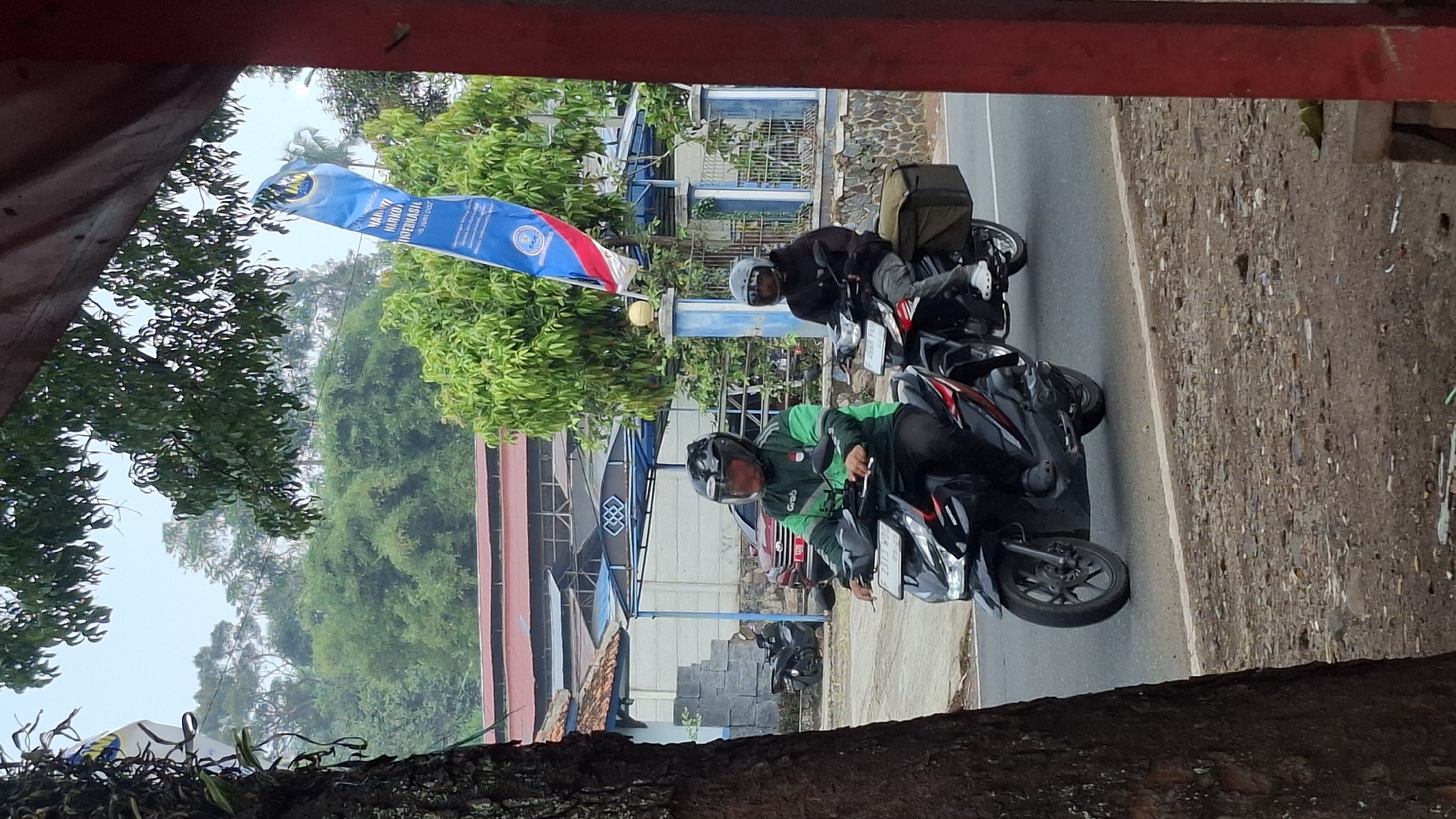 In Cilaku, Indonesia, two individuals are riding motorcycles on a paved road. The motorcycle in the foreground is ridden by a person wearing a green jacket and a black helmet, with the "Grab" logo visible on the jacket. The motorcycle is black. Behind this, another person is riding a black motorcycle, wearing a black jacket and a grey helmet, with a large olive-green bag attached to the rear of the motorcycle. In the background, there are buildings with red roofs and a blue awning, surrounded by lush green trees. A tall, blue banner with text and a logo is unfurled from a pole to the right of the scene. The ground on the left side of the road is covered in dirt and gravel, with a large tree trunk visible. The sky is overcast.
