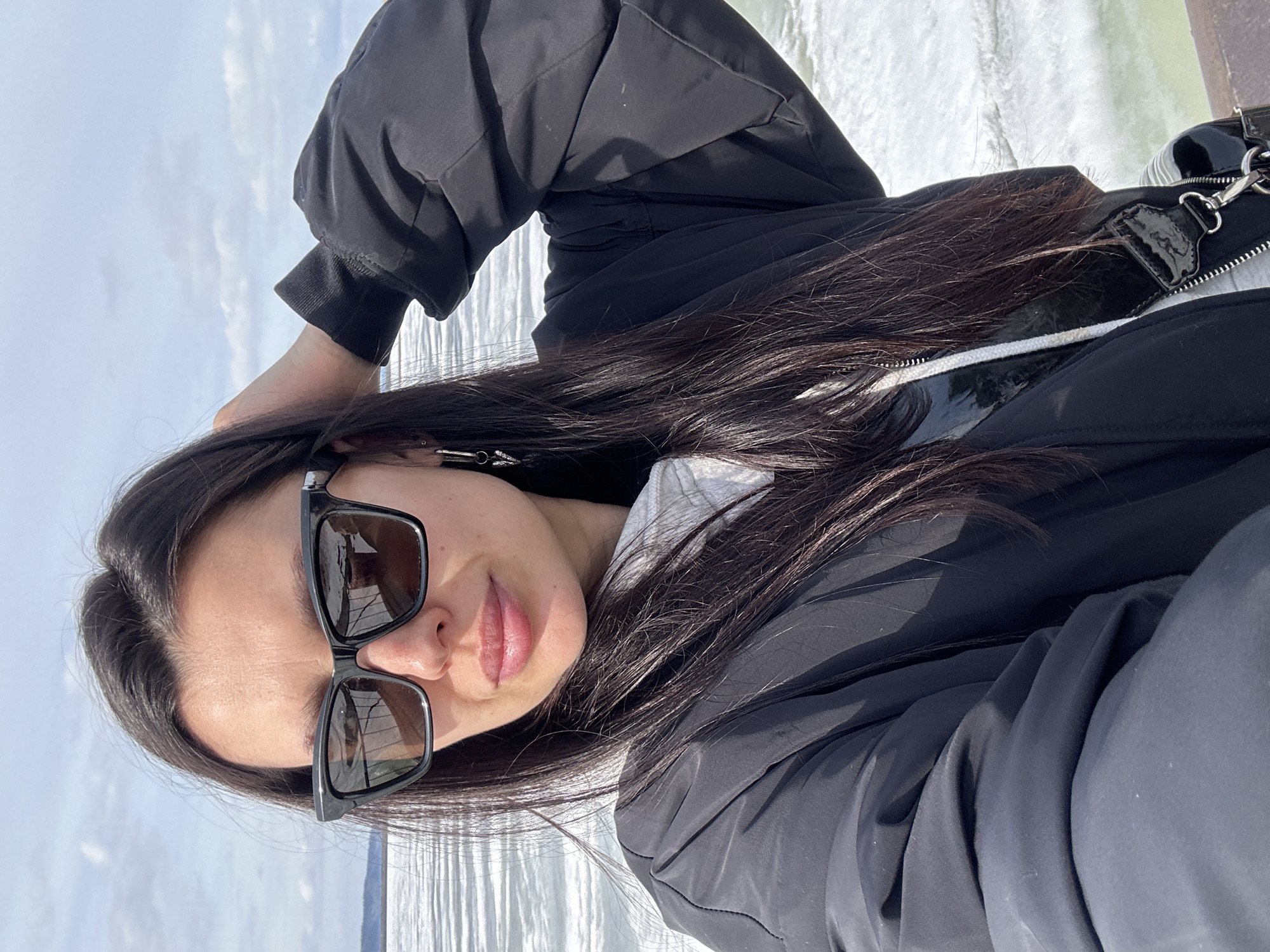 A woman with long, dark hair and sunglasses poses for a selfie. She wears a black jacket over a white hoodie and a black purse strap is visible. The background shows a body of water, likely a river, with reflections of light on the surface. The location is Chaikovsky, Russia.