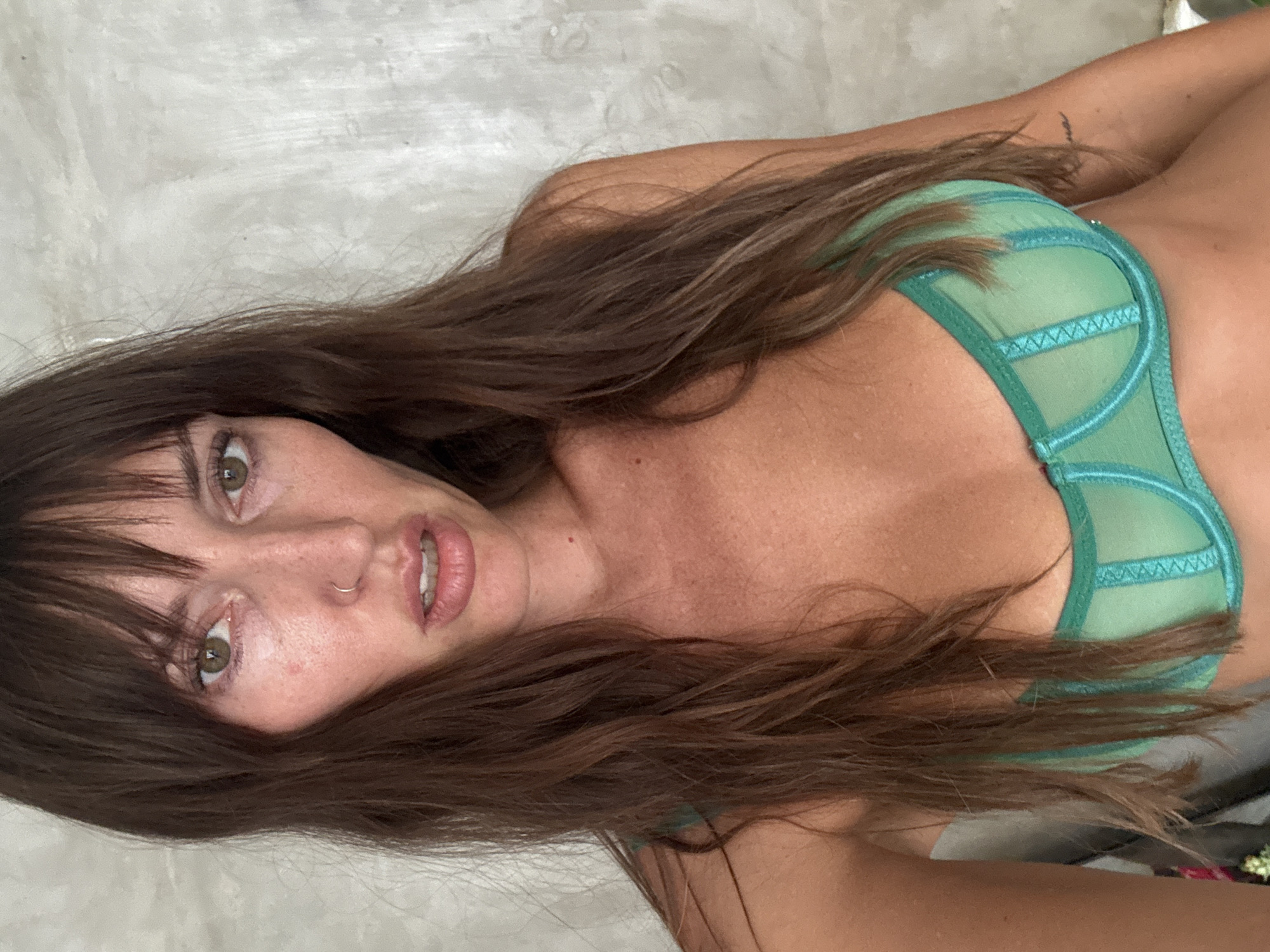 A woman with long brown hair and bangs is lying down, facing the camera, wearing a light green bra. She has a nose ring. The location, North Kuta, Indonesia, cannot be determined from the image alone.