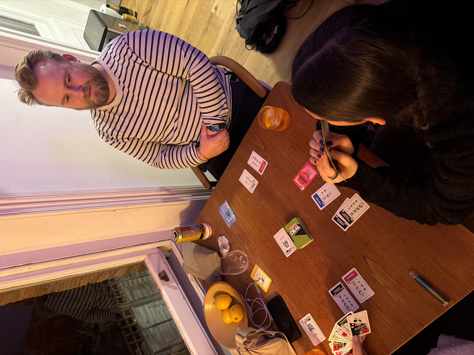 The image depicts an indoor scene within a residential or casual common area in the city of London, United Kingdom, illuminated by warm artificial light. Three individuals are engaged in playing the Monopoly Deal card game around a wooden rectangular table.
In the mid-right, a male subject with fair hair and a beard is seated, wearing a white and navy horizontally striped long-sleeved shirt. He looks towards the table with an attentive expression, his hands clasped, and an Apple Watch visible on his left wrist.
In the foreground, a female subject is viewed from behind, her dark hair styled with a middle part. She holds a fan of Monopoly Deal cards in her hands. Her fingernails are painted dark blue/purple, and she wears a dark ring on her left ring finger. She is clad in a dark, possibly textured, long-sleeved garment. To the far left, the hands of a third individual are visible, also holding game cards.
The wooden table displays the Monopoly Deal game box and numerous game cards arranged in various piles, including money, property, and action cards. Additional items on the table include an open yellow and orange beverage can resting on a white plastic coaster, a glass containing ice and an amber liquid, an empty clear wine glass, a yellow ceramic bowl holding three lemons, a grey fabric pouch, a black mobile phone, a white charging cable, and a small green cylindrical item.
A white-framed window on the left reflects interior elements, including the male subject's striped shirt, and a dark, blurry exterior environment. The interior walls are white, with a pink/purple light source illuminating the wall section adjacent to the window. In the background right, a grey waste bin and several bottles are visible on a light wooden floor.