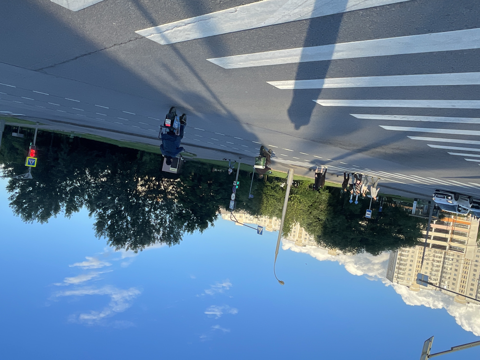 The image depicts a street scene in Moscow, Russia, likely during the daytime. A pedestrian crossing is prominent, marked by white stripes on the asphalt road. On the left side of the road, a traffic light shows a red signal. A few people are visible, some crossing the street and others standing on the sidewalk. One person is riding a scooter or a similar electric vehicle. Trees line the street, and in the background, several multi-story residential buildings are visible. The sky is blue with some scattered white clouds. The overall impression is of a typical urban environment. The shadows suggest it's daytime, and the clear sky indicates fair weather. There are no strong emotional cues. Some text is visible on a license plate of a scooter, appearing to read "390 YAK." Other signs are present but not clearly legible.