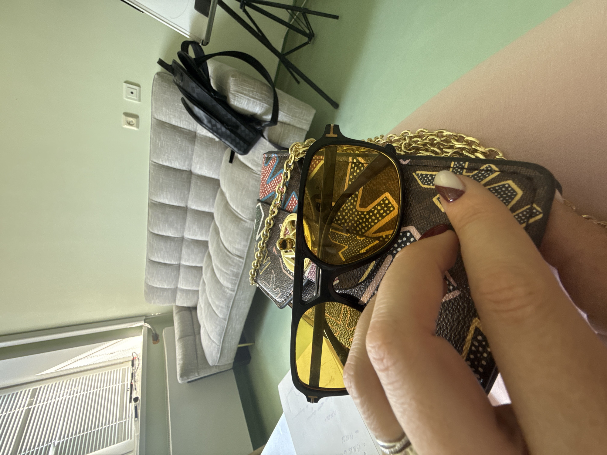 The image displays an indoor scene, likely a residential room in Moscow, Russia, captured from a slightly elevated, tilted perspective. In the foreground, a person's hands are holding a pair of sunglasses with reflective yellow lenses and a black frame, which features a small 'T' logo above the bridge. The sunglasses rest on a small, dark brown patterned purse or wallet with a gold chain strap. The purse's pattern consists of abstract geometric shapes and stylized letters that resemble 'M', 'A', 'X', or 'V'. The person's fingers are adorned with a silver ring, and their nails are manicured with a two-tone design, featuring white and a dark red/brown polish.
The background reveals a light green wall where two white electrical sockets or switches are mounted. To the right, a segment of a light grey upholstered sofa or headboard with a tufted, rectangular pattern is visible. A black bag rests on top of this furniture. Further back, parts of a black tripod stand can be seen. To the left, a window with horizontal blinds allows bright daylight to illuminate the room, indicating it is daytime. Below the sunglasses, a piece of white paper with faint, largely indecipherable text and numbers, including what appears to be "10 000", is present. The scene depicts a casual, everyday moment, with no specific emotions discernible.
