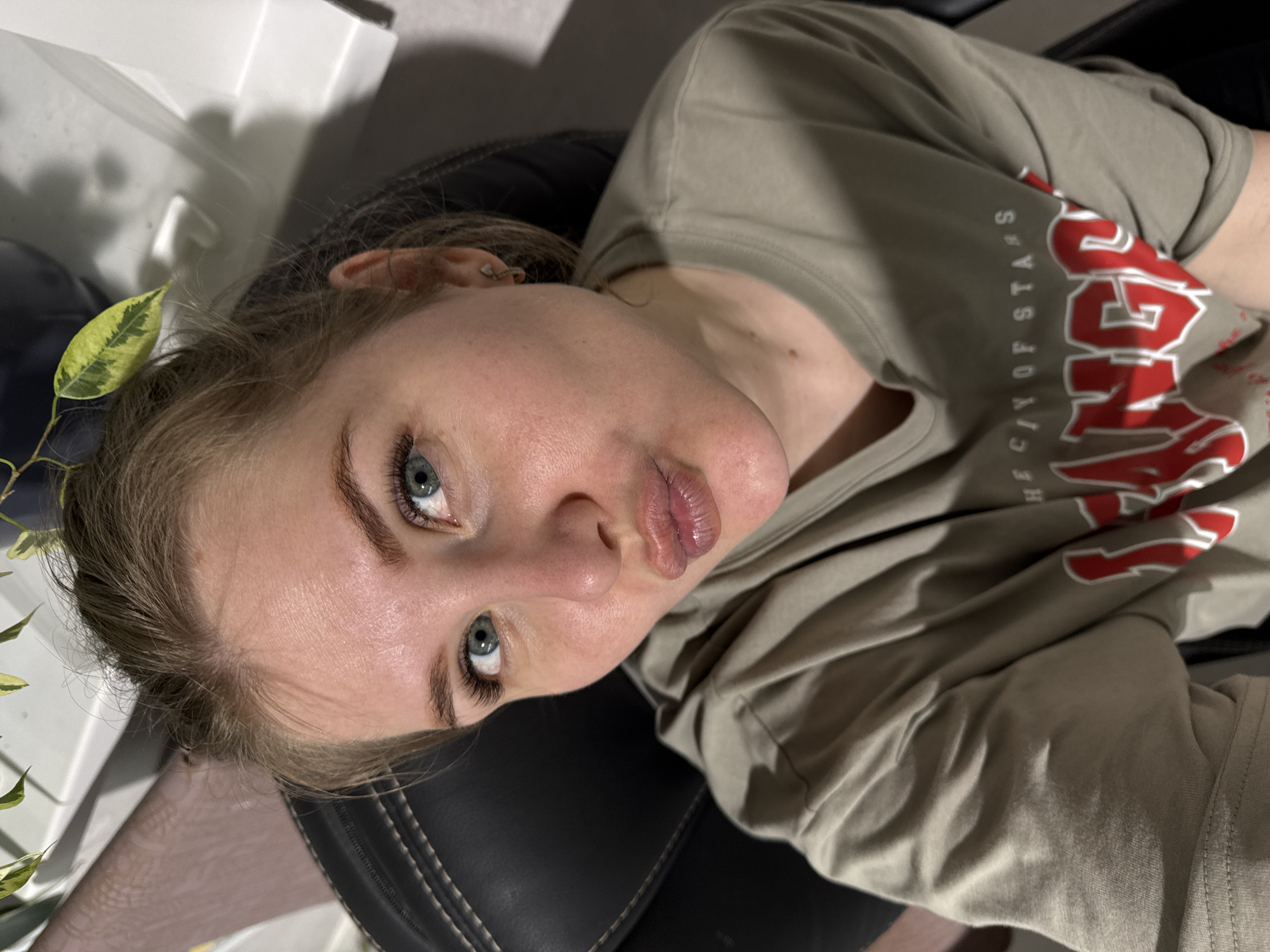 A woman with light skin and blue eyes is lying back in a black chair. She is wearing a light brown t-shirt with the word "Vegas" in red and white, and the phrase "The City of Stars" in white printed on it. She is looking directly at the camera with slightly pursed lips. A plant with variegated green and yellow leaves is near the left side of her head. A white surface is visible in the upper left corner of the image. The lighting is somewhat dim. The location cannot be determined from the image.