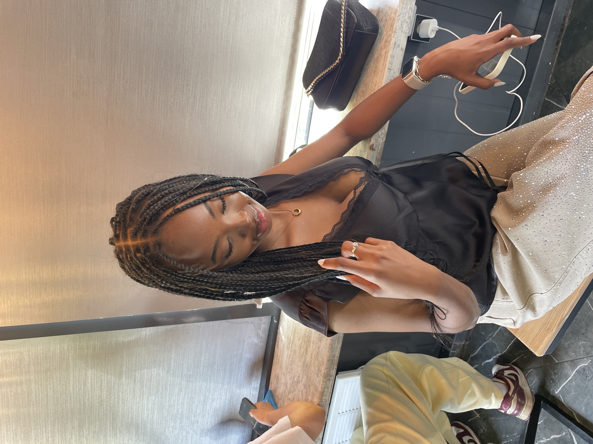 A young Black woman with braided hair and fair skin is seated indoors, looking down and to her right. She wears a black satin top with lace trim and a gold necklace with a circular pendant. Her left hand rests on her thigh, and her right hand is raised, touching her hair and holding a ring. She also wears a silver watch on her right wrist and a ring on her left index finger. Her pants are tan and covered in small, sparkling embellishments. To her right, a black handbag with a gold chain strap rests on a counter. To her left, another person wearing a light pink shirt and light yellow pants is visible, holding a smartphone. The background consists of a textured wall and window blinds. The overall setting appears to be a cafe or a similar casual establishment.