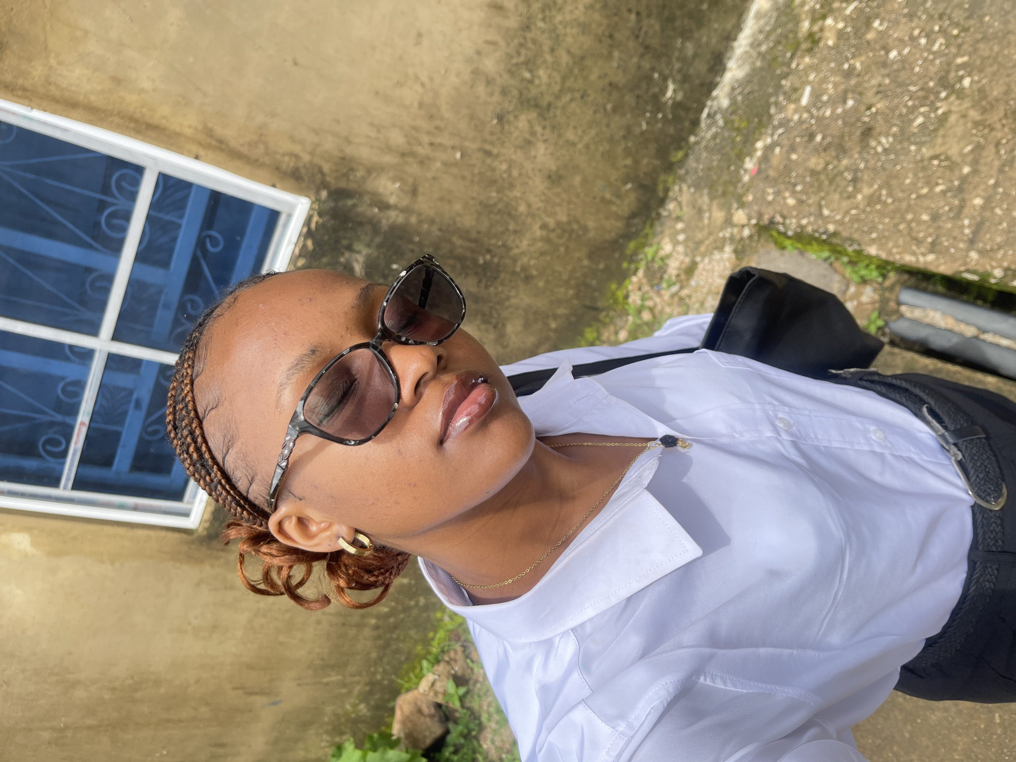 A young Black woman stands outdoors, taking a selfie. She is wearing a white button-down shirt, black pants, sunglasses, and gold hoop earrings. A delicate gold chain with a dark pendant rests on her neck. Her hair is braided and styled in a curly updo. She is carrying a black bag. The background consists of a textured, tan-colored wall with a blue-grated window. The ground is a rough, concrete surface with some green vegetation. The scene appears to be in a low-income urban or semi-urban setting, consistent with areas in Rafin Par, Nigeria.
