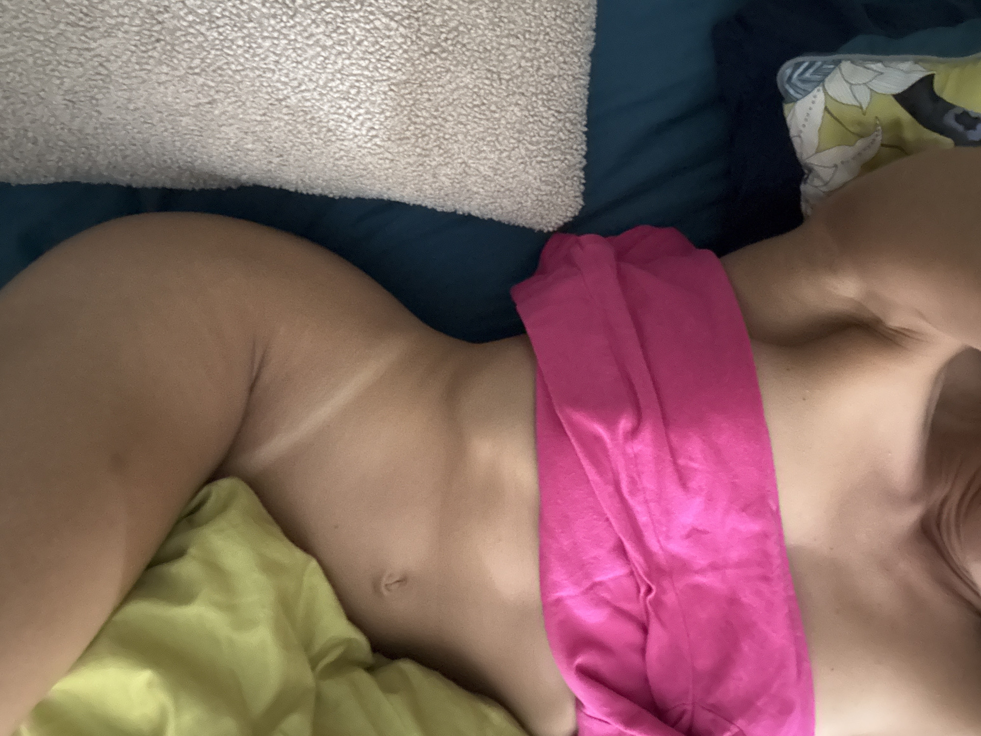 The media file is a close-up, overhead shot of a person lying down. The person's torso, from the side of their chest down to their hip, is visible. They are lying on a bed with a blue sheet. A light-colored, textured pillow is at the top of the frame, and a brightly colored floral pillow is to the right. A section of lime green fabric is visible under the person's hip, and a draped piece of bright pink fabric covers their abdomen. The person's skin tone is light, and there are visible tan lines on their side. The lighting suggests it is daytime, and no specific weather cues are apparent. The setting appears to be a bedroom. No text is visible in the image.
