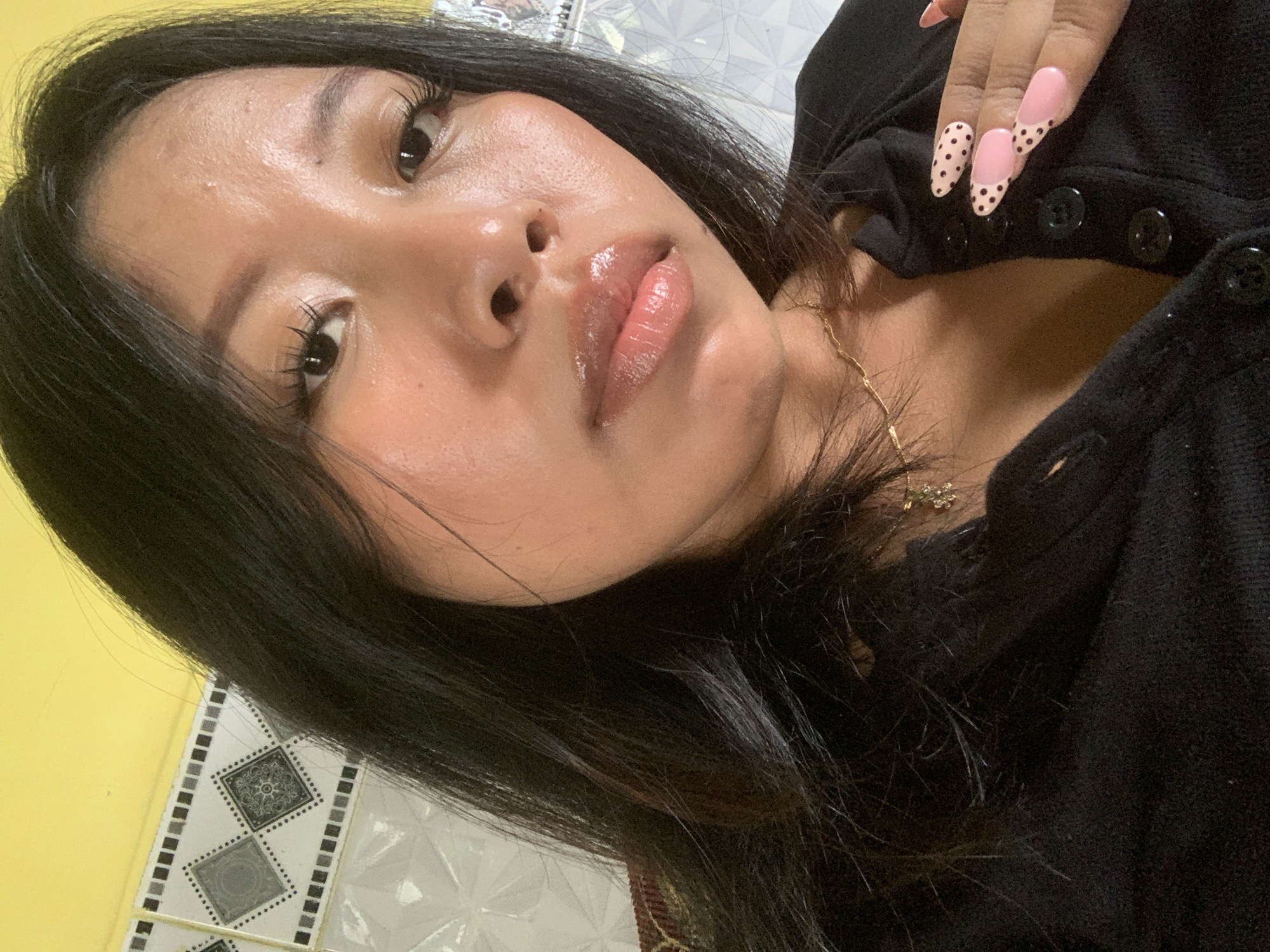 A young woman with dark hair, wearing a black top, poses for a selfie indoors. She has on long, light-pink fingernails with black polka dots and French tips. She is wearing a delicate gold necklace. The background consists of yellow and tiled walls. The photo appears to have been taken in Jajag, Indonesia.
