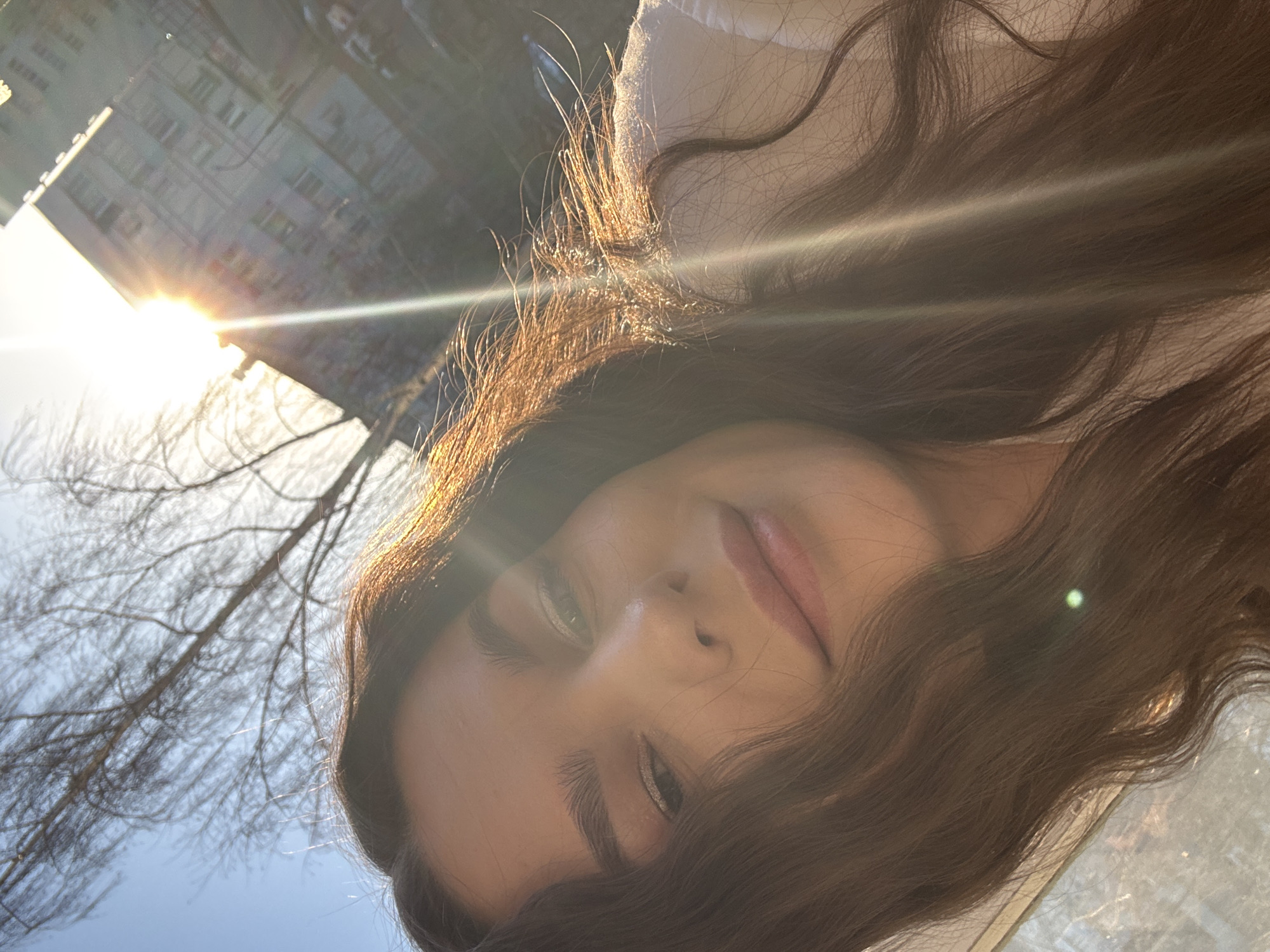 A low-angle, close-up photograph shows a young woman's face, angled toward the camera, with long brown hair framing her face. Sunlight shines from the upper left, creating a lens flare and illuminating the tips of her hair. Behind her, in the background, are bare tree branches silhouetted against a bright sky. Partially visible behind the branches is a multi-story building. Based on the cityscape and information provided, the image is likely taken outdoors in Chaikovsky, Russia, though specific details such as street names or landmarks are not visible.