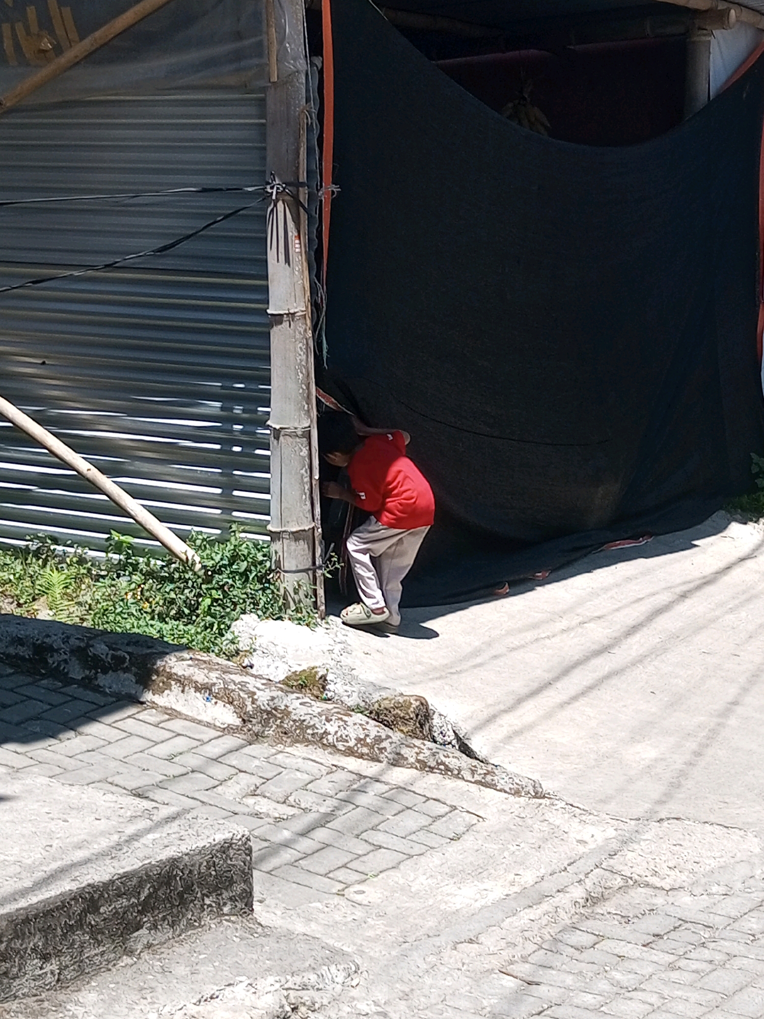 An individual wearing a red long-sleeved shirt and light-colored pants is stooping down next to a concrete pole, partially obscured by a large black tarp. The person appears to be reaching or looking into something behind the tarp. In the background, a corrugated metal wall is visible, along with some greenery. The ground is paved with concrete and there are some stone elements. The scene is outdoors, under bright sunlight, casting shadows on the ground. This location is identified as Andongsili, Indonesia.