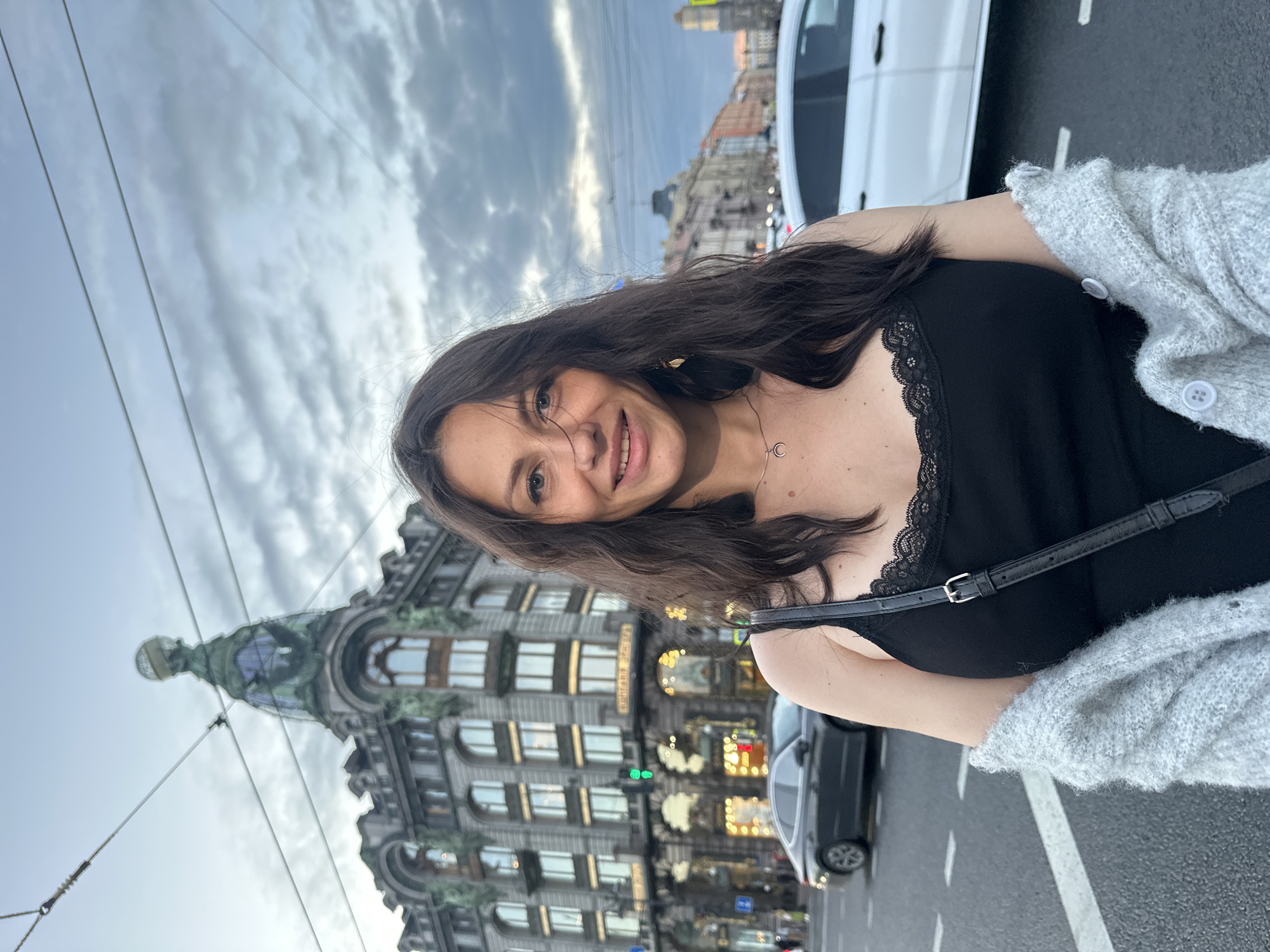 A young woman with long, dark hair is standing on a city street in Saint Petersburg, Russia. She is wearing a black camisole with lace trim, a gray cardigan draped over her shoulders, and a black crossbody bag. She is smiling and looking at the camera. In the background, there is a grand, ornate building with a distinctive dome topped by a globe. Cars are visible on the street, and overhead wires suggest the presence of public transportation. The sky is overcast with dramatic clouds, indicating either early morning or late evening. The architecture of the building points to it being a historic landmark, possibly the Singer House (Dom Knigi) given its unique dome and location on Nevsky Prospekt.