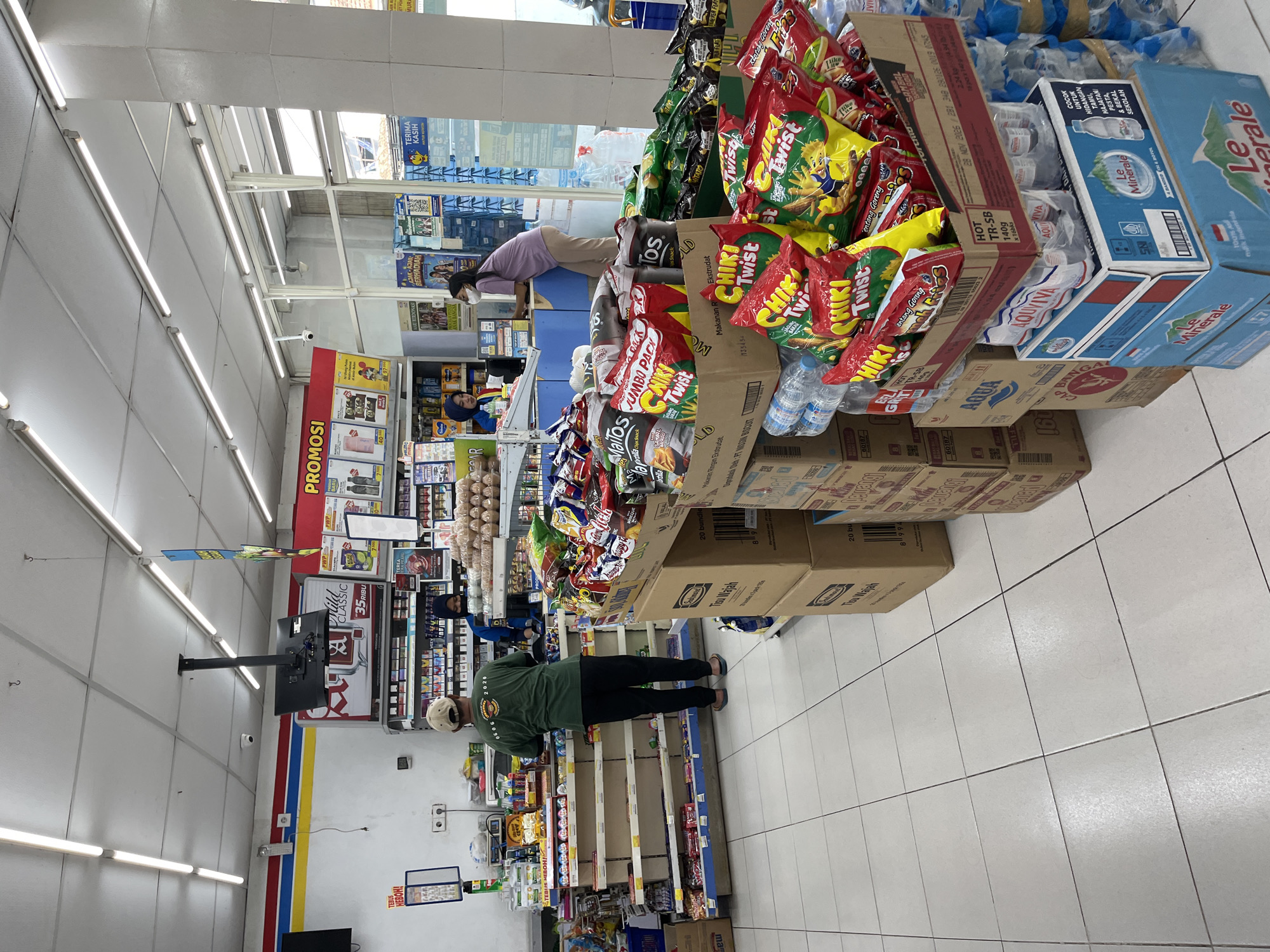 The image shows the interior of a convenience store in Kedensari, Indonesia. The store is well-stocked with various products, including snacks, drinks, and toiletries, displayed on shelves and in boxes. Two women are visible, one wearing a blue uniform and standing behind a counter, possibly attending to a customer or stocking shelves. The other woman, wearing a light purple shirt and beige pants, is interacting with a computer at the counter. A man in a green shirt and beige cap is standing in front of a shelf, facing away from the camera. The lighting is bright, typical of a retail environment, with fluorescent lights illuminating the tiled floor and ceiling. Promotional posters and signage are visible on the walls and shelves. The overall impression is of a bustling, organized retail space.