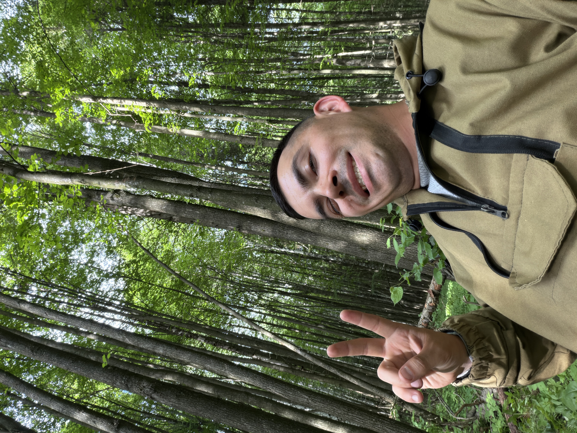 An upward-angle selfie-style portrait captures a male individual in a dense forest, located in Kechur, Russia. The man, appearing to be in his late 20s to 40s, is positioned in the lower center of the frame. He has short dark hair, a light complexion, and is looking directly at the camera with a broad smile, showing teeth, and a slight squint in his right eye. He is wearing a khaki or olive-drab colored anorak featuring a half-zip front, a hood with a drawstring, and black zipper details; a grey shirt is visible beneath. His left hand is raised to head height, making a peace sign (V-sign). The background is dominated by a dense growth of numerous tall, slender trees with greyish-brown bark and vibrant green foliage, indicating a lush forest environment. Sunlight filters through the tree canopy, illuminating the leaves and creating bright patches. Green undergrowth and some fallen branches are visible on the forest floor in the lower sections of the image.