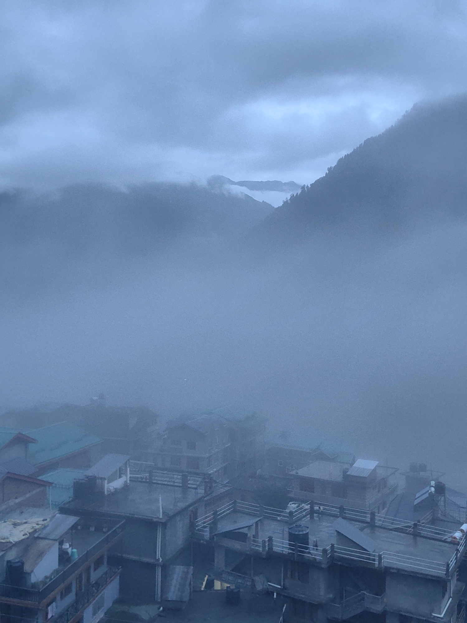 The image depicts a misty, overcast scene in the town of Vashisht, India. In the foreground, several multi-story buildings with flat roofs are visible, some with railings and water tanks. The roofs are mostly dark, with some showing lighter colored materials or exposed pipes. One building in the lower left has balconies with decorative railings and a large black water tank. Further back, more buildings are visible, some with pitched roofs, including one with a teal-colored roof. The middle ground and background are dominated by thick fog and mist, obscuring the lower slopes of a mountain range. The mountain slopes are covered in trees, with the upper portions disappearing into the clouds. The overall atmosphere is somber and atmospheric, suggesting a cool, damp day. No people are clearly discernible in the image, and no actions or interactions are captured.