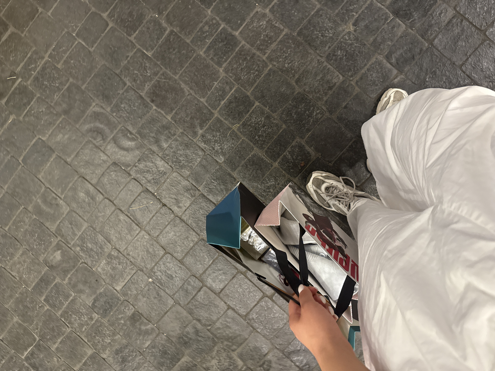 A person's hand holds two shopping bags filled with items. The bags are patterned with red and white images. The person is wearing white athletic shoes and white traditional clothing. The ground is paved with dark, rectangular stones. The scene is outdoors, likely on a street or walkway. No specific location in Yerevan, Armenia is identifiable from the image.