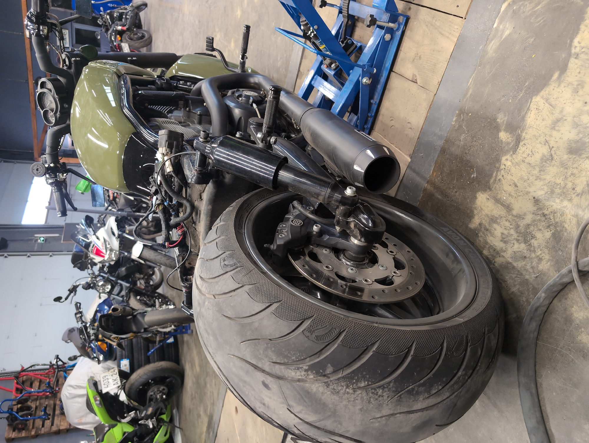 An olive green motorcycle is parked indoors, possibly in a garage or workshop. The motorcycle is the primary subject, positioned in the foreground and taking up a significant portion of the frame. It is missing some body panels and appears to be undergoing maintenance or repair.
The front of the motorcycle, including the handlebars, fuel tank, and engine area, is visible. The engine is exposed, with wiring and mechanical components readily apparent. The fuel tank is a prominent olive green color with a glossy finish. The handlebars are black and equipped with various controls.
The rear of the motorcycle features a large, wide tire with a distinct tread pattern. A portion of the brake disc and caliper assembly is visible. A black exhaust pipe, large in diameter, is mounted on the right side of the motorcycle. The rear suspension components, specifically shock absorbers, are also visible.
In the background, other motorcycles are present, including a bright green sportbike on the left and several other bikes in various states of repair or storage. The environment appears to be a workshop or storage area, with tools and equipment visible in the background, including a blue hydraulic lift supporting the olive green motorcycle. The floor is concrete, with some scuff marks and debris. The lighting suggests an indoor environment with artificial light sources. The image is taken from a low angle, emphasizing the size and presence of the motorcycle.
