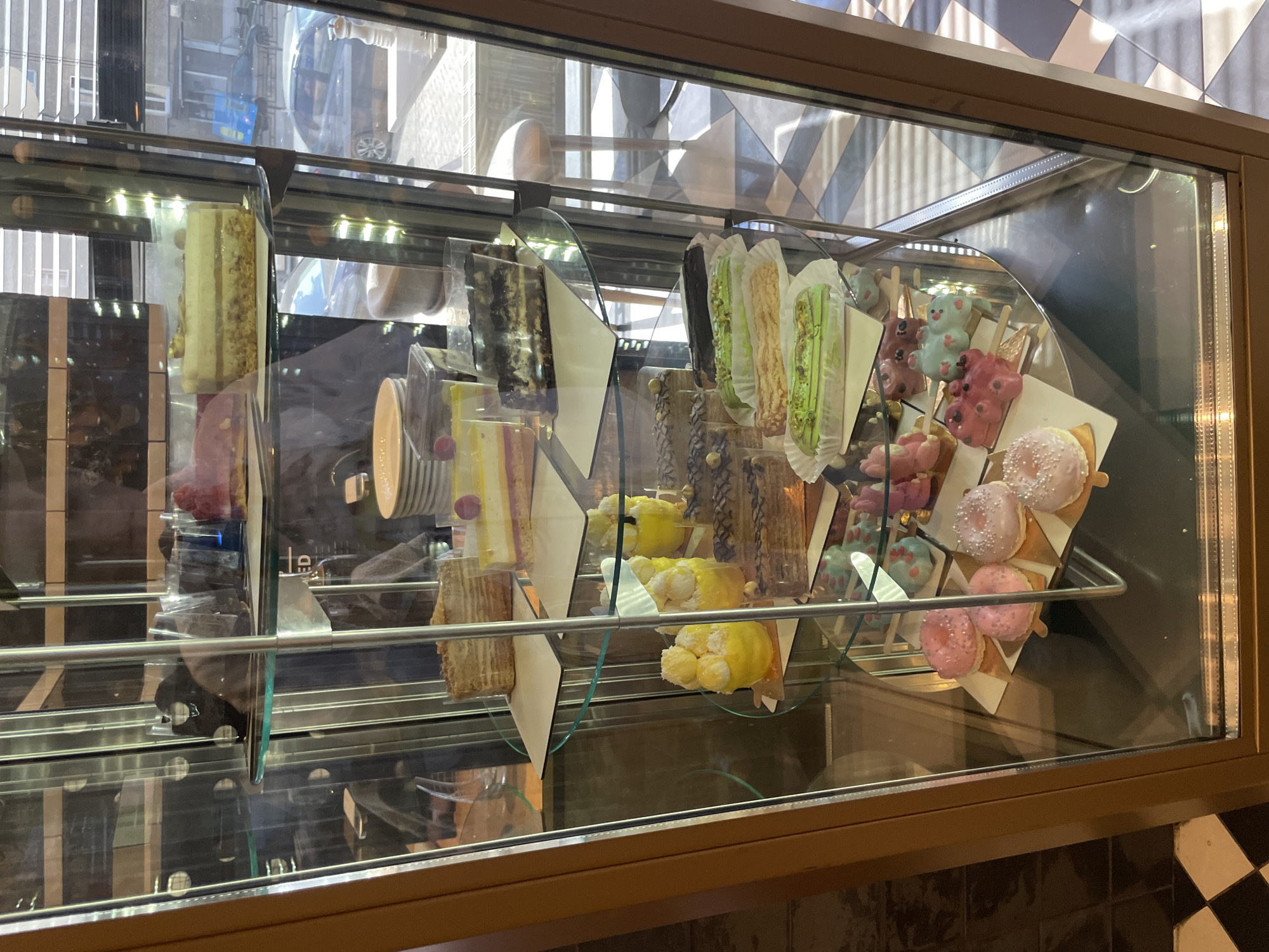 The image shows a refrigerated display case filled with a variety of desserts. The desserts include cake slices, eclairs, teddy bear-shaped treats on sticks, and pink frosted donuts on sticks. The treats are displayed on small paper stands within the glass case. The background visible through the glass includes reflections of a city street, possibly in Izhevsk, Russia, and the interior of the building where the display case is located. The overall impression is of a confectionery or cafe showcasing its offerings.