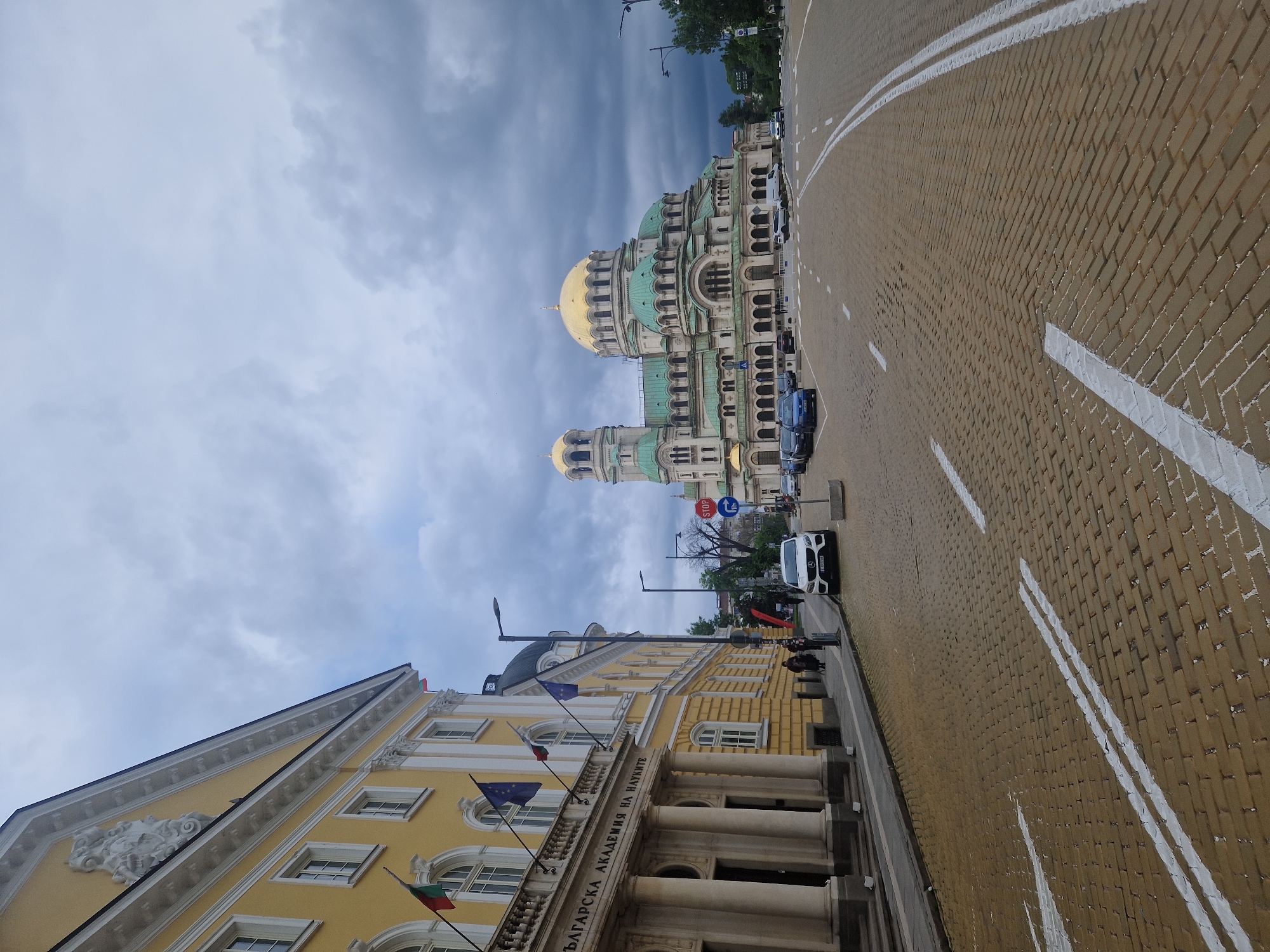 The image shows a street scene in Sofia, Bulgaria. On the right side of the frame is a wide cobblestone street marked with white lines. Several cars are parked along the edge. In the background is the Alexander Nevsky Cathedral, notable for its golden domes and elaborate green and white architecture. To the left, a yellow building is visible, identified by its inscription in Bulgarian as the Bulgarian Academy of Sciences. Flags are flying from the building's facade. The sky is overcast with dark, cloudy skies.