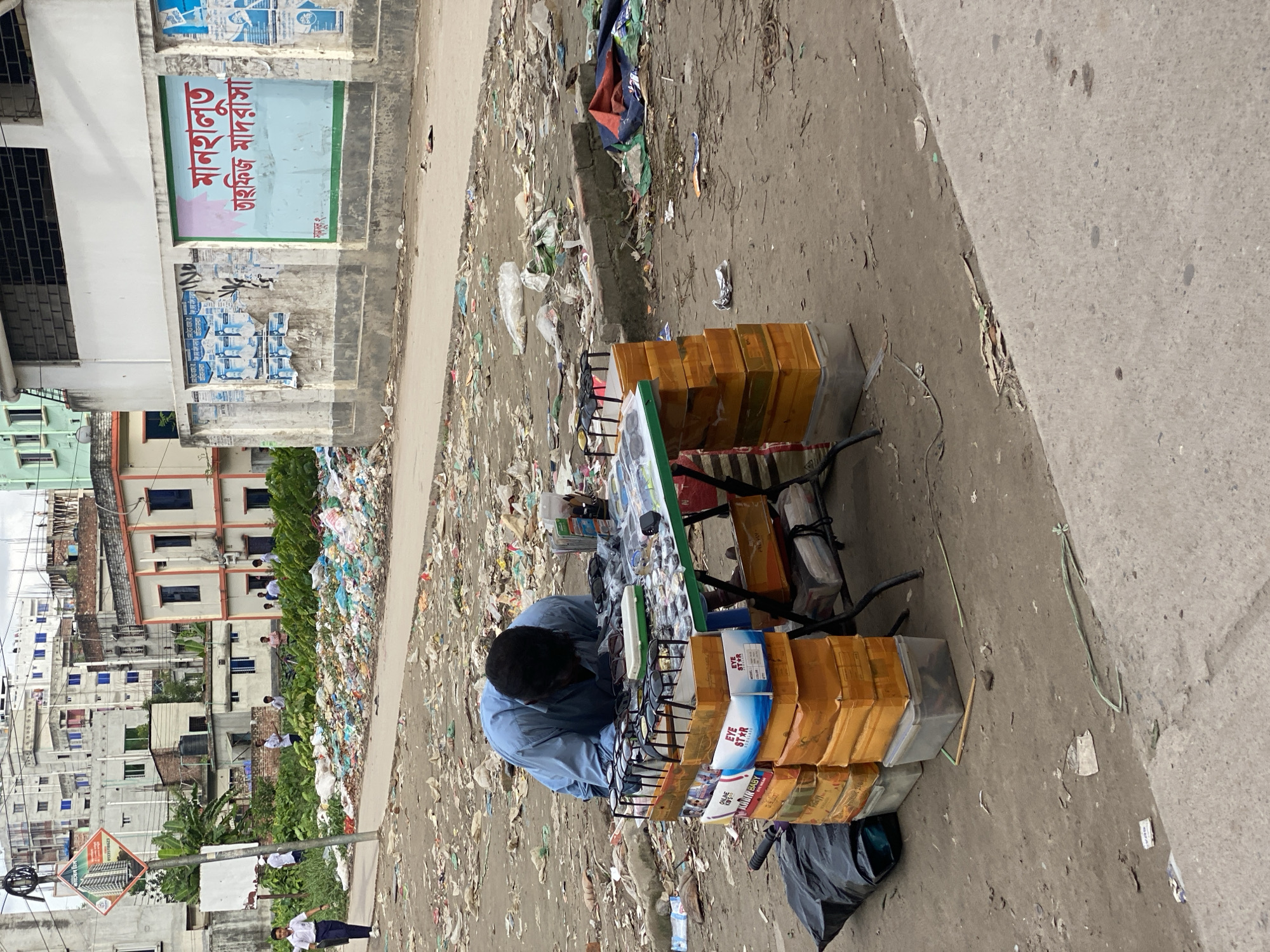 In an outdoor scene in Dhaka, Bangladesh, a vendor is seen hunched over a table displaying numerous pairs of sunglasses. The table is laden with sunglasses in various styles and colors, neatly arranged on stands and in boxes. The vendor, dressed in a light blue shirt, is focused on his merchandise. The ground surrounding the table is littered with trash, including plastic bags, paper, and debris, indicating a rather unkempt environment. In the background, several buildings are visible, suggesting an urban setting. The overall impression is one of a busy street market with a prevailing atmosphere of clutter and everyday commerce.