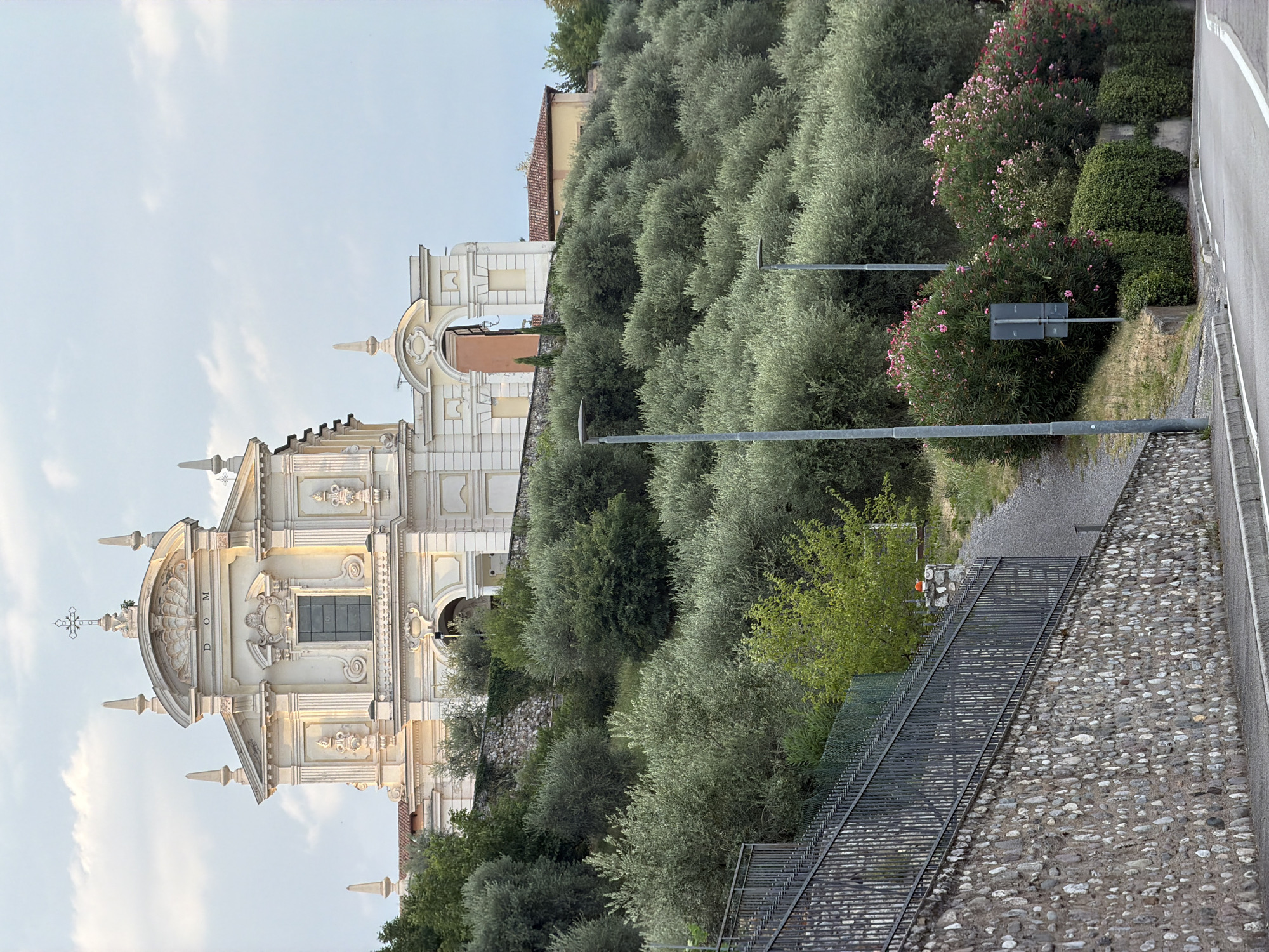 The image shows the Church of Santa Maria Assunta in Polpenazze del Garda, Italy. The church is situated on a hilltop, overlooking a landscape filled with olive trees and lush greenery. The facade of the church is ornate, featuring classical architectural elements, a prominent cross on its roof, and the inscription "D OM". The sky above is partially cloudy with a soft, diffused light, suggesting it might be late afternoon or early morning. In the foreground, a stone wall and a metal fence line a road that curves upwards towards the church. Several streetlights are visible along the road and the hillside. The overall scene conveys a sense of serene beauty and historical significance.
