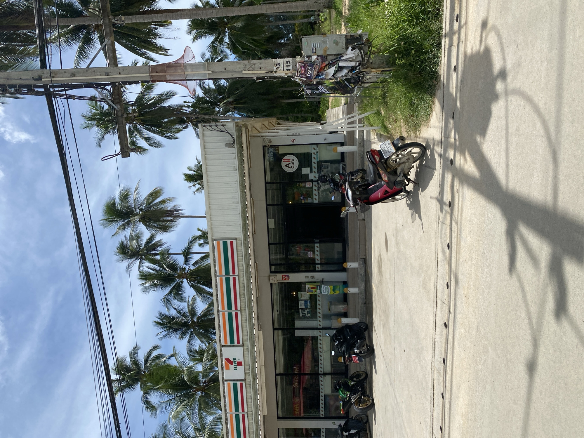A 7-Eleven convenience store is situated on a street in Tambon Koh Phangan, Thailand, under a bright, partially cloudy sky. The store features a white, corrugated metal roof and a glass facade with a prominent "7-Eleven" logo displayed above. To the right of the entrance, an "All Cafe" sign is visible. Several motorcycles are parked in front of the store on the concrete pavement, including a green motorcycle, a black motorcycle, and a red and black scooter. A utility pole with numerous wires and posters is positioned to the right of the store, adjacent to a patch of green grass and vegetation. Palm trees are scattered throughout the background, suggesting a tropical environment. The street appears to be paved with concrete, with a visible drain grate and shadows cast by overhead wires and vegetation on the ground. The scene is captured from a slightly elevated perspective, looking towards the store.