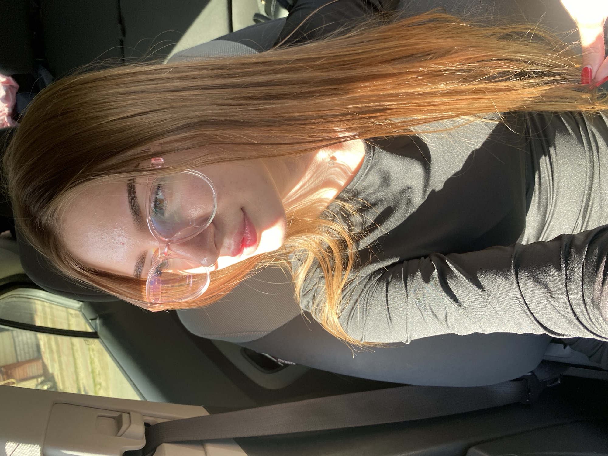 The media file shows a close-up, angled selfie of a young woman inside a vehicle. The woman has long, straight, auburn hair that is illuminated by sunlight, creating highlights. She is wearing clear-rimmed glasses with a pink tint and a black long-sleeved shirt. Her left hand, with bright red nail polish, is visible near her hair on the right side of the frame. Sunlight is casting a shadow and creating a bright glare on her face and neck.
The background indicates the setting is inside a car, with parts of the car's interior visible, including a seat and a seatbelt. Through the car window, there is a glimpse of an outdoor scene with greenery and what appears to be a building or fence, suggesting daytime. The lighting suggests it is a sunny day, possibly late afternoon or early morning due to the angle of the light. The overall mood is relaxed and casual. No visible text is present in the image. The location context provided is Дьяково, Russia.