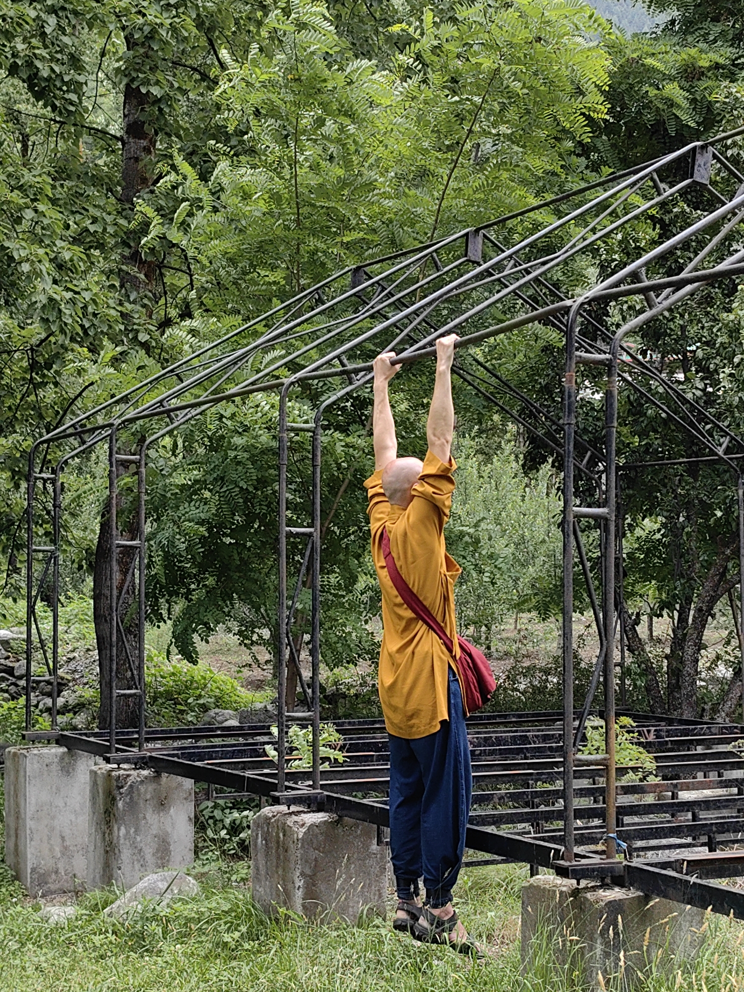 A bald, male figure, wearing a saffron robe and dark blue pants, hangs from a metal structure in Vashisht, India. The structure appears to be an unfinished or derelict frame, possibly for a greenhouse or a similar outdoor installation, supported by concrete blocks. He is viewed from behind, with his arms fully extended, gripping a horizontal bar. A red bag is slung over his right shoulder. The background consists of lush green foliage, suggesting a natural or semi-rural environment within the town.