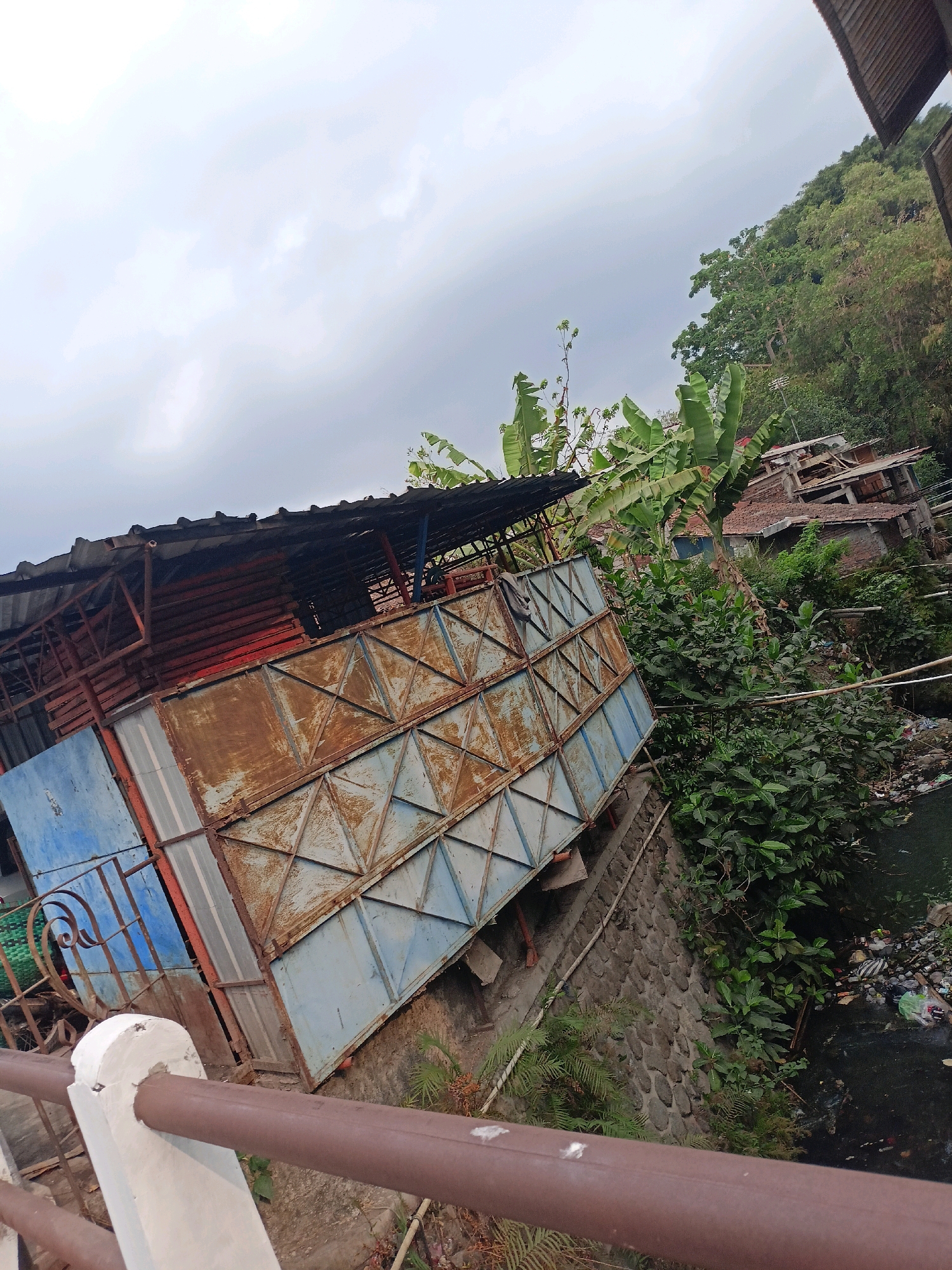 The image depicts a section of the city of Bandongan, Indonesia, characterized by its rural setting. A weathered, multi-colored corrugated metal structure dominates the foreground, its surfaces exhibiting rust and peeling paint. The structure appears to be a makeshift building or storage area, with a corrugated metal roof supported by a metal frame. A brown metal railing runs along the bottom of the frame, indicating a walkway or elevated platform.
Adjacent to the metal structure is a waterway, its surface choked with an accumulation of plastic waste and debris. The banks of the waterway are lined with a stone retaining wall, overgrown with lush green vegetation. Behind the structure and the waterway, a steep, densely forested hillside rises, dotted with a few scattered buildings. Banana trees with their large, distinctive leaves are visible among the foliage, suggesting a tropical climate. The sky is overcast with heavy, grey clouds, hinting at impending rain. There are no people visible in the image. The scene conveys a sense of neglect and environmental concern, with the natural beauty of the landscape marred by the presence of pollution.