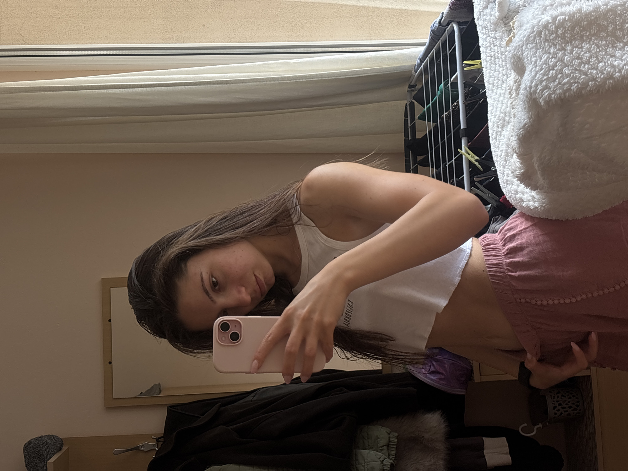 A young woman with long brown hair takes a mirror selfie in what appears to be a bedroom. She is wearing a white crop top and pink shorts. Her phone, with a pink case, is held up to capture the reflection. The background shows a closet rack with clothes and a window with white curtains. A white fluffy blanket is draped over the closet rack. The lighting suggests it is daytime.