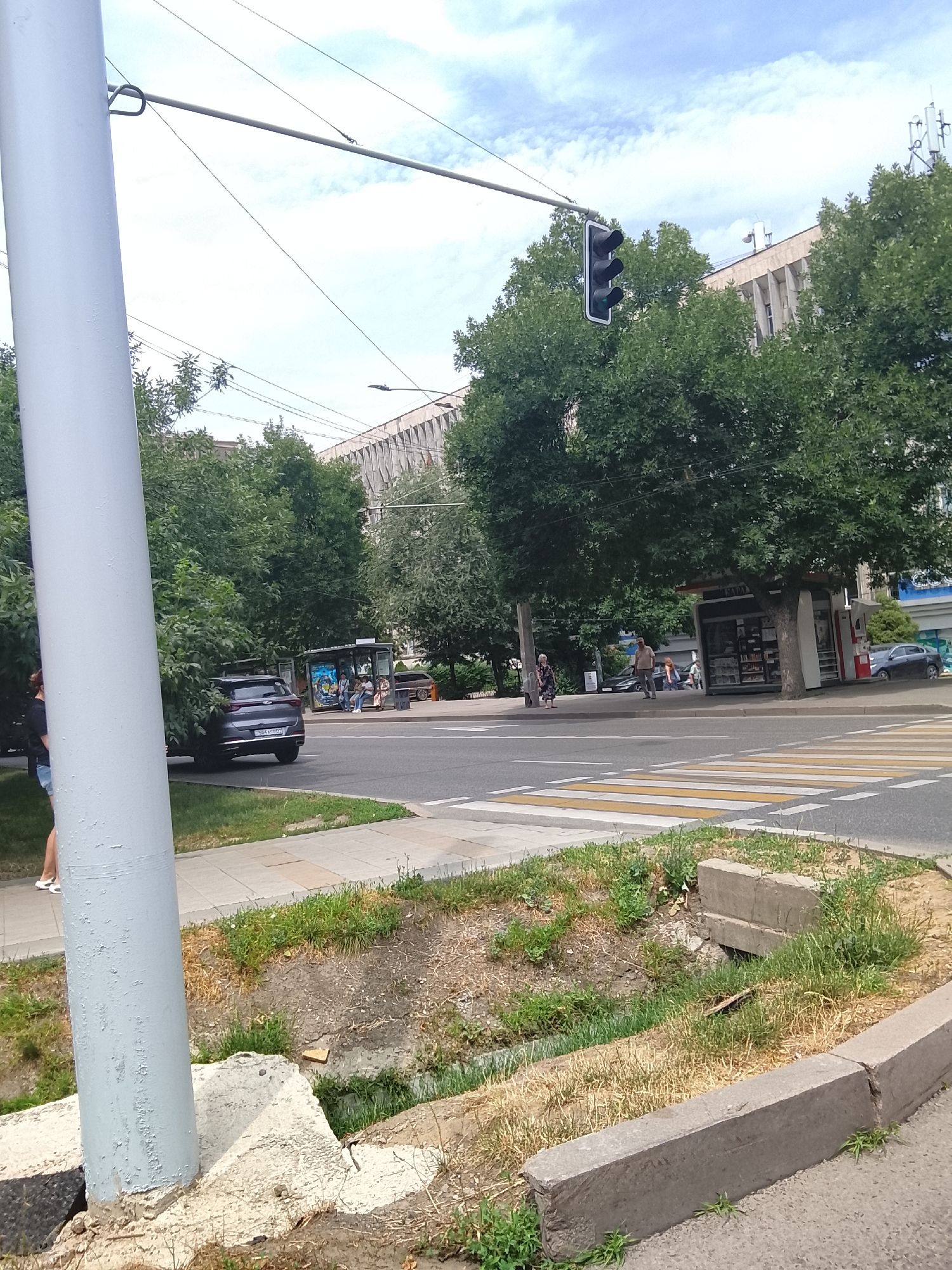 A wide shot captures a street scene in Almaty, Kazakhstan. In the foreground, a grey utility pole stands prominently on the left, with dry grass and a concrete curb bordering a ditch on the right. A pedestrian crosswalk with yellow and white stripes stretches across the road. A dark-colored SUV is parked on the right side of the road, partially obscured by trees. A bus stop shelter with people seated inside is visible further down the street. Trees line the sidewalks and form a canopy over parts of the road. Traffic lights hang over the intersection. In the background, a large, multi-story building with a patterned facade is visible, along with utility poles and wires against a cloudy sky.