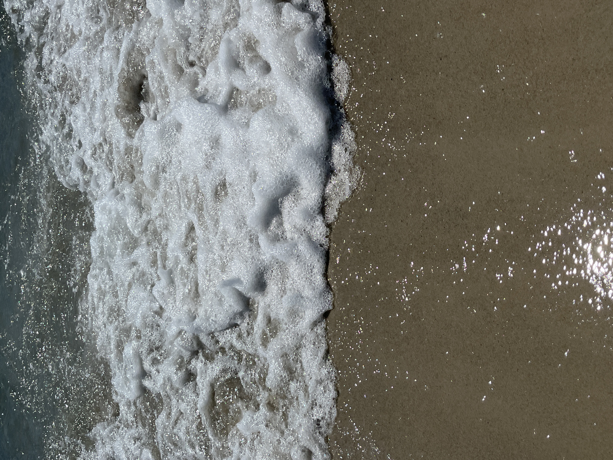 The image shows a close-up, overhead view of a sandy beach with the edge of a wave lapping onto the shore. The sand is a light brown color and appears wet, with small sparkles of light reflecting on its surface. The water is foamy and white, forming a frothy edge where it meets the sand. The top portion of the image shows the darker, murkier water of the ocean with small whitecaps and sparkles of light. The scene is tranquil and focuses on the natural elements of the beach. There are no people or man-made objects visible in the image.