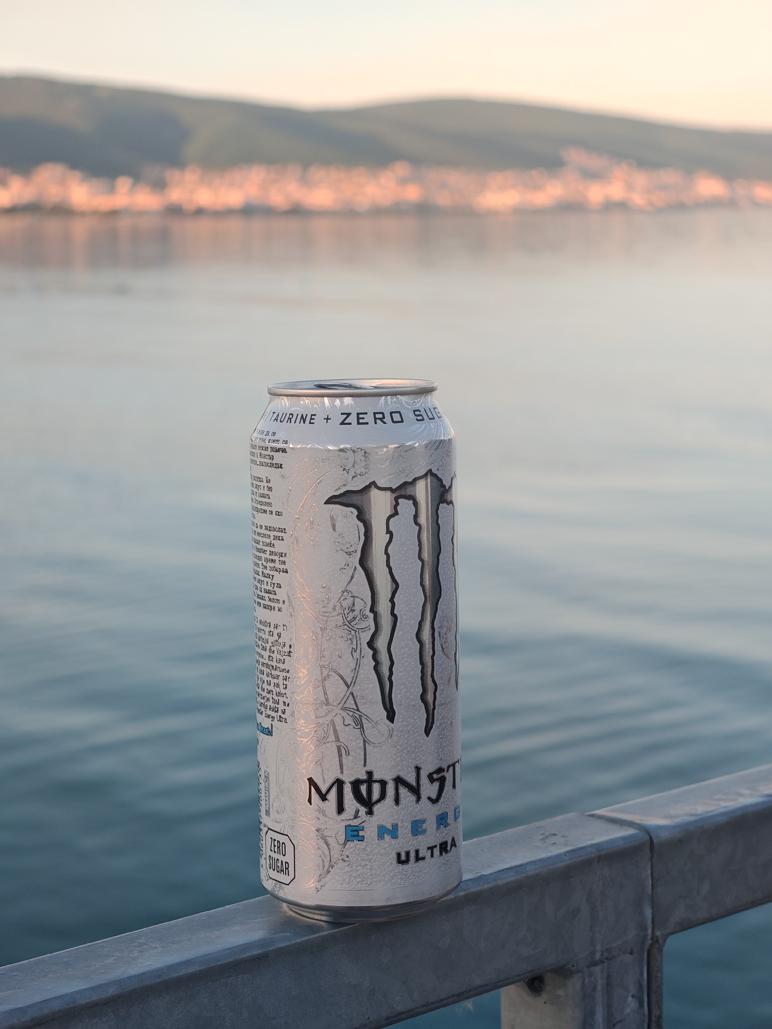 The image shows a can of Monster Energy Ultra drink, specifically the white, zero-sugar variety. It is positioned on a metal railing, likely near a body of water. In the background, there is a blurred cityscape or town along the shoreline of the body of water. The overall setting appears to be outdoors during daylight, possibly at dawn or dusk, given the soft lighting.