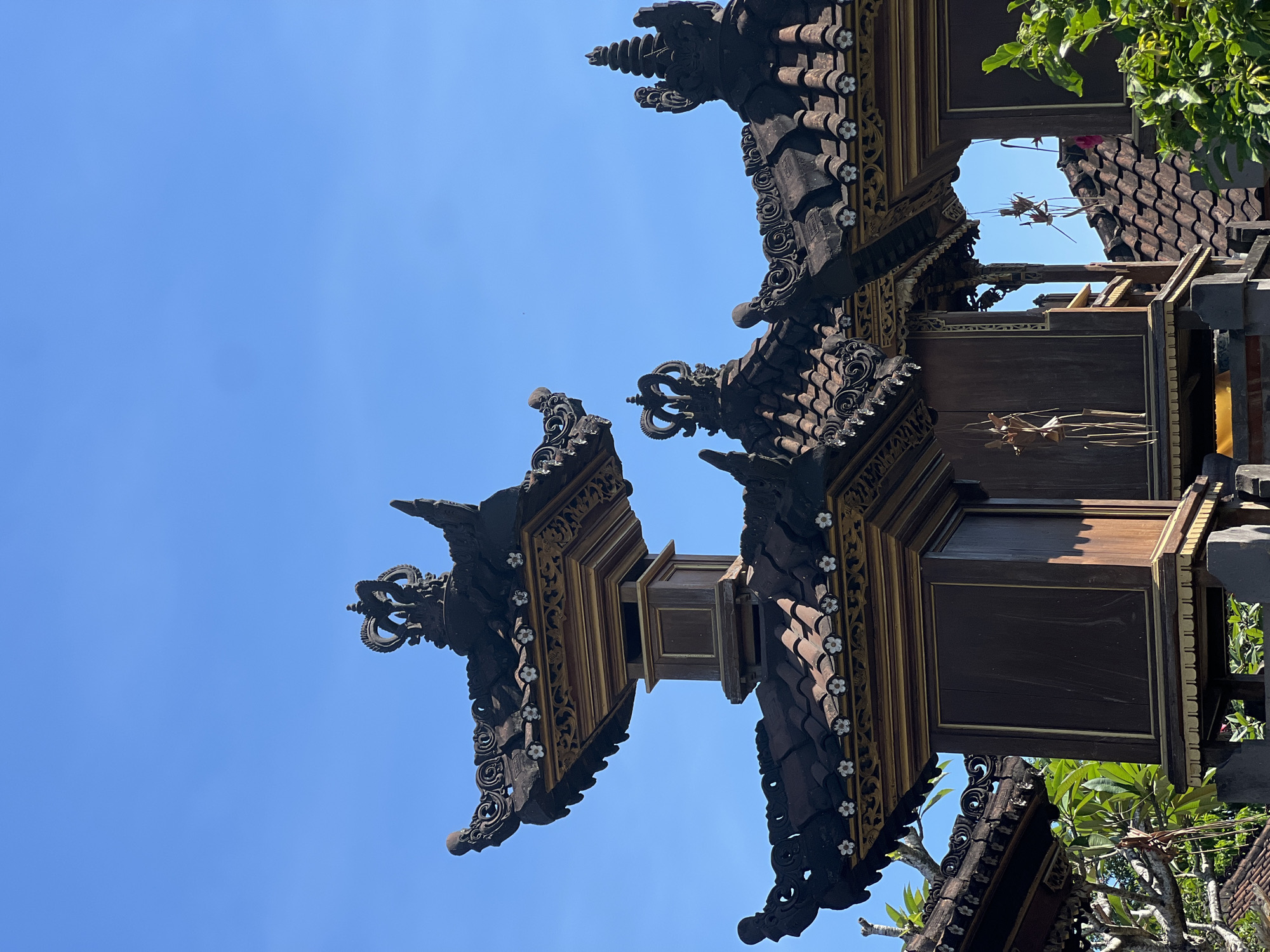 The image shows the ornate, multi-tiered roof structures of a Balinese temple against a clear blue sky. The roofs are made of dark, tiled material and adorned with intricate dark brown carvings, gold trim, and small white flower-like decorations. Several decorative crowns are visible atop the highest points of the structures. Wooden pillars and panels form the supporting structures. In the foreground and background, lush green foliage is visible. Some delicate, dried-leaf ornaments hang from the eaves of one of the structures. The architecture is characteristic of temples found in Sukawati, Indonesia, a region known for its traditional crafts and culture. No people are present in the frame.