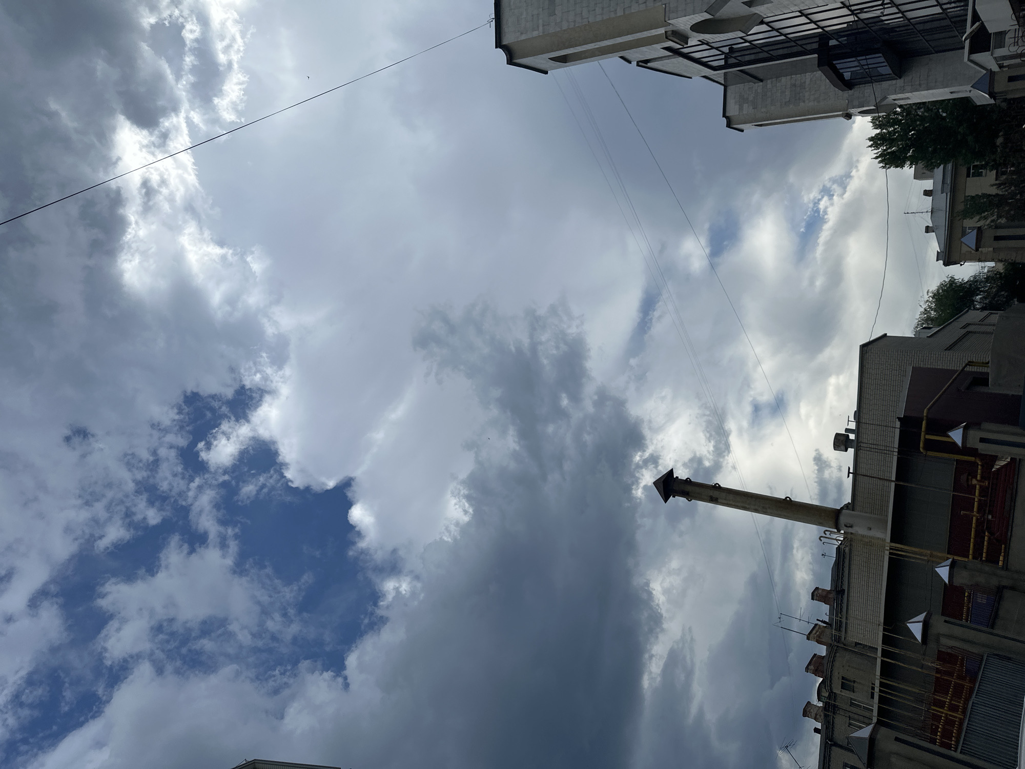 The image captures a view of the sky above the city of Saratov, Russia, with some buildings and structures visible in the foreground. The sky is partly cloudy, with both dark and light clouds present. A tall, narrow chimney, likely from an industrial building, stands prominently on the left side of the frame, leaning slightly. To the right, a modern building with large windows and an arched facade is visible. Several utility wires are stretched across the sky. The scene appears to be an outdoor urban environment under a variable sky.
